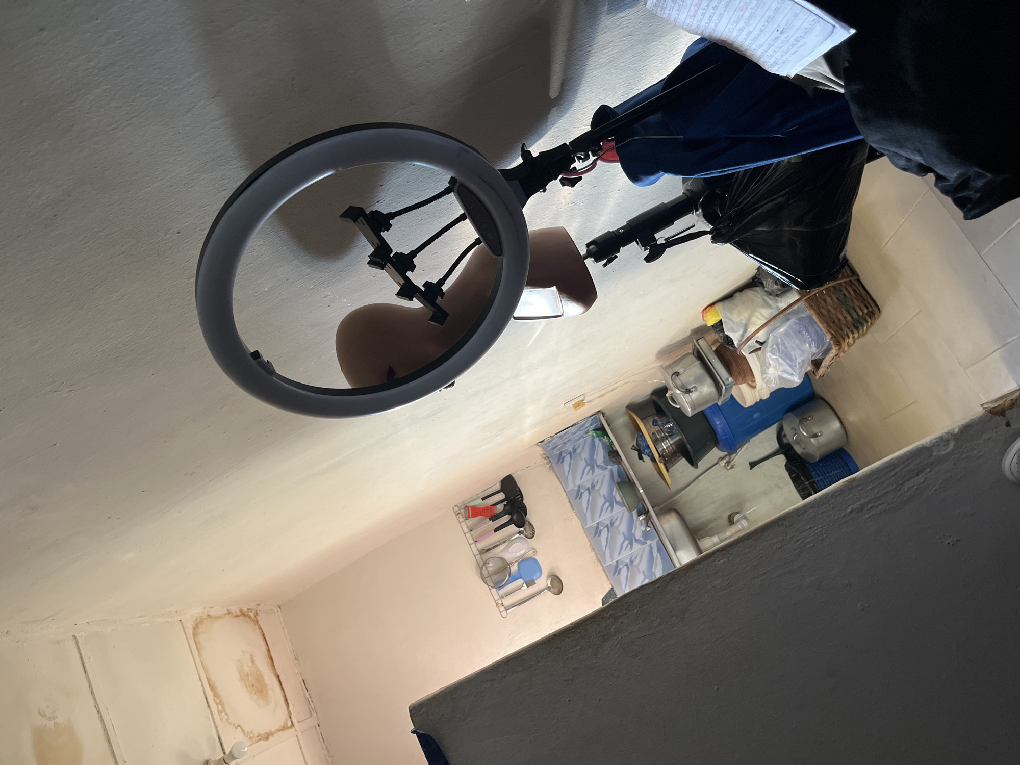 This is a forensic, matter-of-fact description of a scene, potentially located in Jos, Nigeria. The image depicts an indoor space that appears to be a kitchen or a utility area. The walls are painted a light color, and the ceiling shows signs of water damage with prominent brown stains. In the foreground, a large, dark gray vertical object, possibly a refrigerator or a tall cabinet, occupies the left side of the frame, partially obscuring the view of the rest of the room.
To the right of this object, a kitchen sink area is visible. Above the sink, a rack holds various kitchen utensils, including ladles, spatulas, and a sieve. The backsplash behind the sink is tiled with a blue and white patterned tile. Various items are on the counter and floor, including pots, buckets, a woven basket, and plastic bags. A large blue bucket is prominent on the floor, and next to it, a black plastic bin.
In the mid-ground, a ring light is set up on a stand, with a mannequin head positioned behind it, suggesting the setup might be for filming or photography. A dark blue garment is draped over the stand. A notebook with handwritten text is visible in the lower right corner of the image. The flooring appears to be tiled. The overall impression is of a functional, somewhat cluttered domestic space.