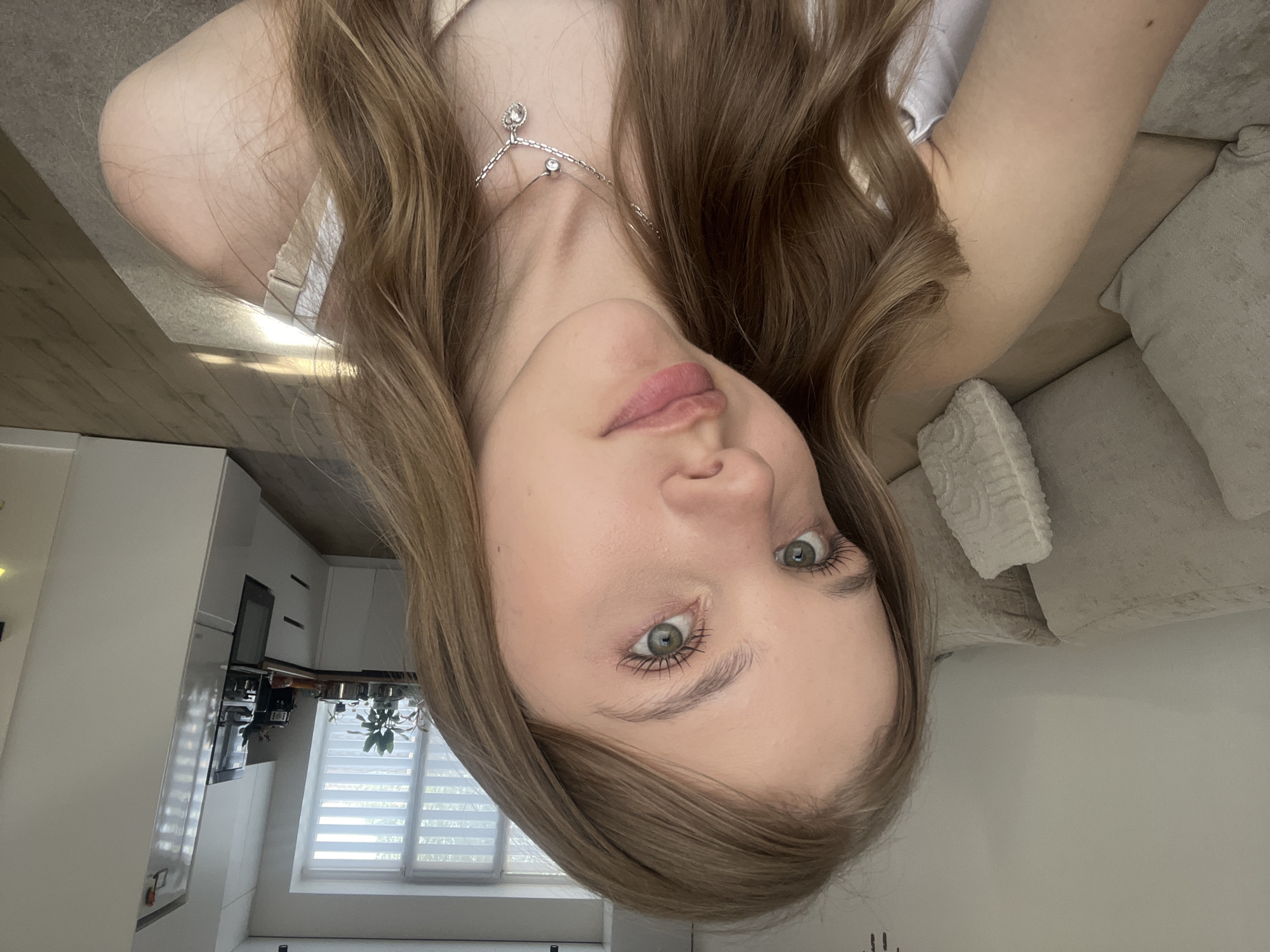 A young woman with light skin, light brown hair, and green eyes is looking directly at the camera. She is wearing a necklace with a small pendant. The background includes a kitchen with white cabinets and appliances, and a window with blinds. To her right, there is a sofa with cushions. The location cannot be determined as Votkinsk, Russia without additional information.