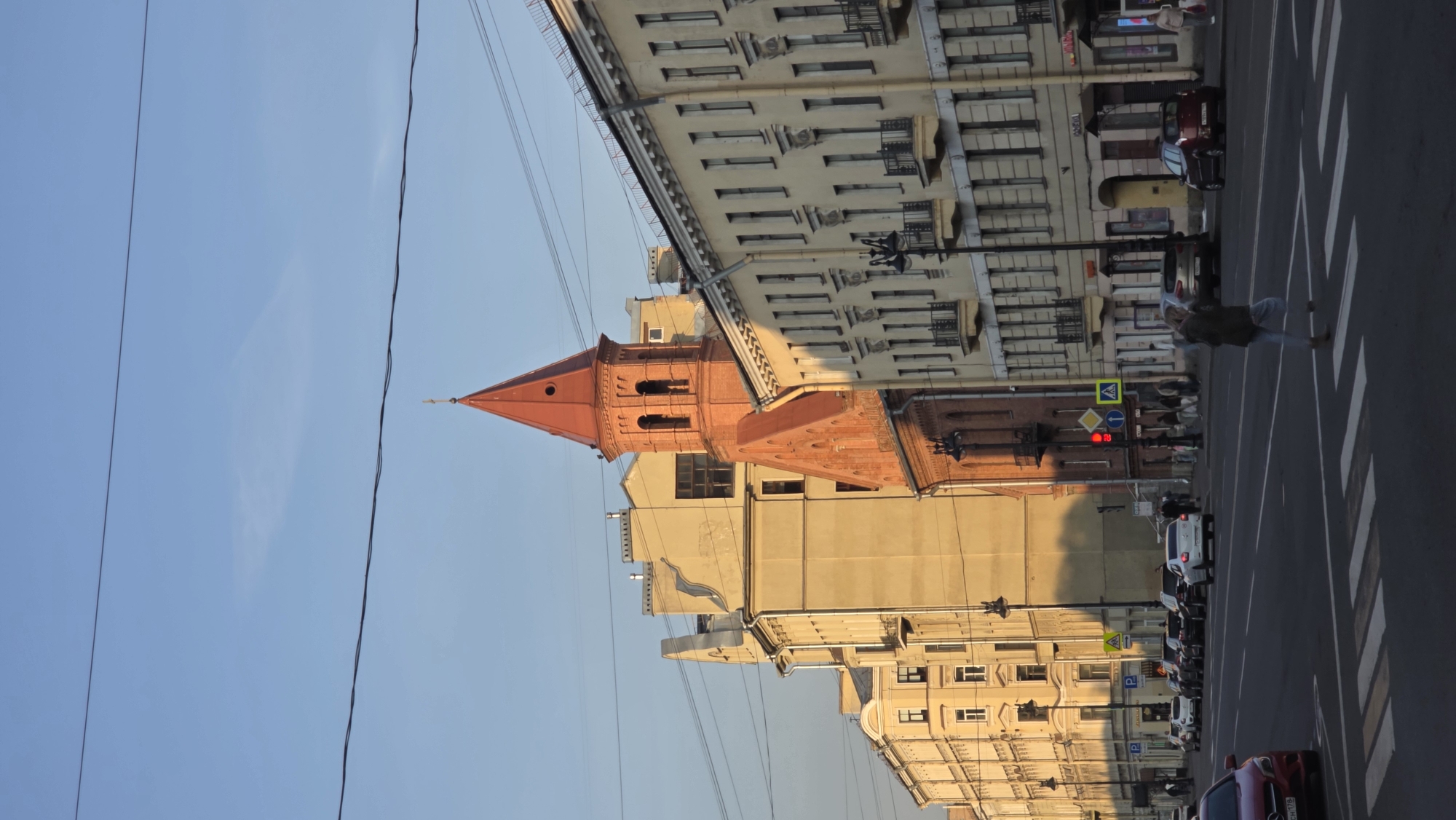 The image shows a street scene in Saint Petersburg, Russia, with the Evangelical Lutheran Church of Saint Anne visible. The church has a distinctive red brick tower with a pointed spire. The building next to the church is light yellow. The street is lined with buildings of various heights and architectural styles. There are cars parked on the street and traffic lights at a pedestrian crossing. There are also people crossing the street. Overhead power lines run across the sky. The sky is blue.