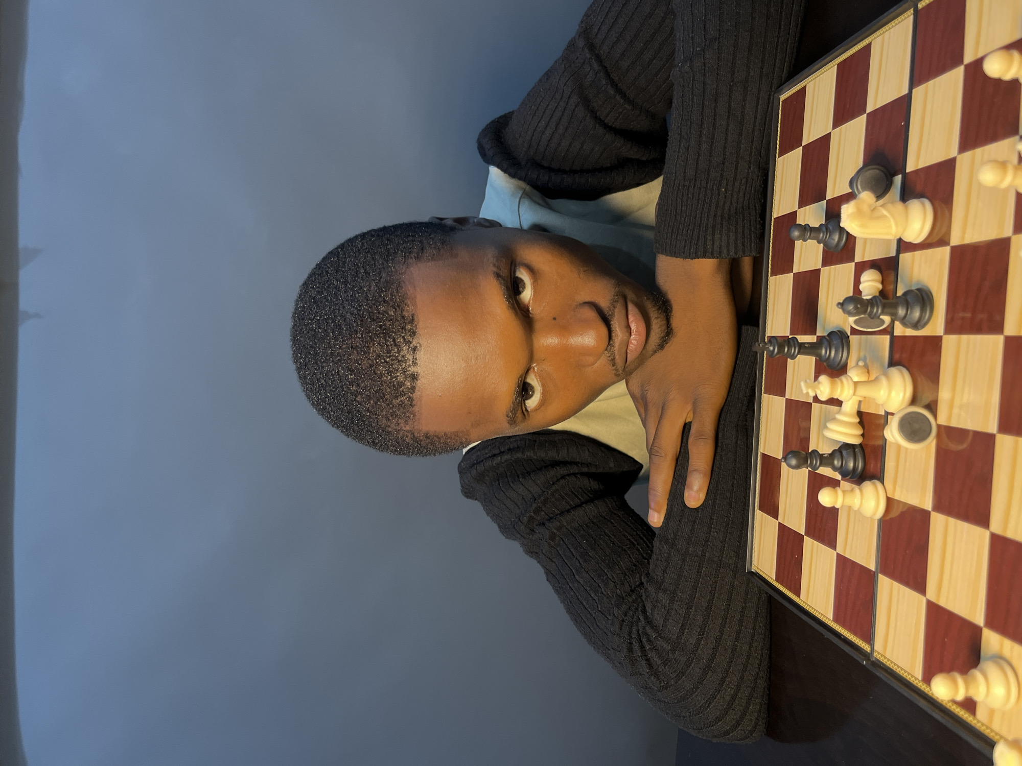 A young Black man with short hair and a beard is leaning over a chessboard with his arms resting on the table. He is looking directly at the camera with a serious expression. The chessboard has several pieces on it, some upright and some knocked over, suggesting an ongoing or recently finished game. The man is wearing a black ribbed sweater over a light-colored vest. The background is a plain, dark wall. The lighting is from the front and slightly to the left, casting shadows on the man's face and the chessboard. The image appears to be taken indoors, possibly in a home or a study. There is no discernible information in the image to indicate the location as Gura Top, Nigeria.