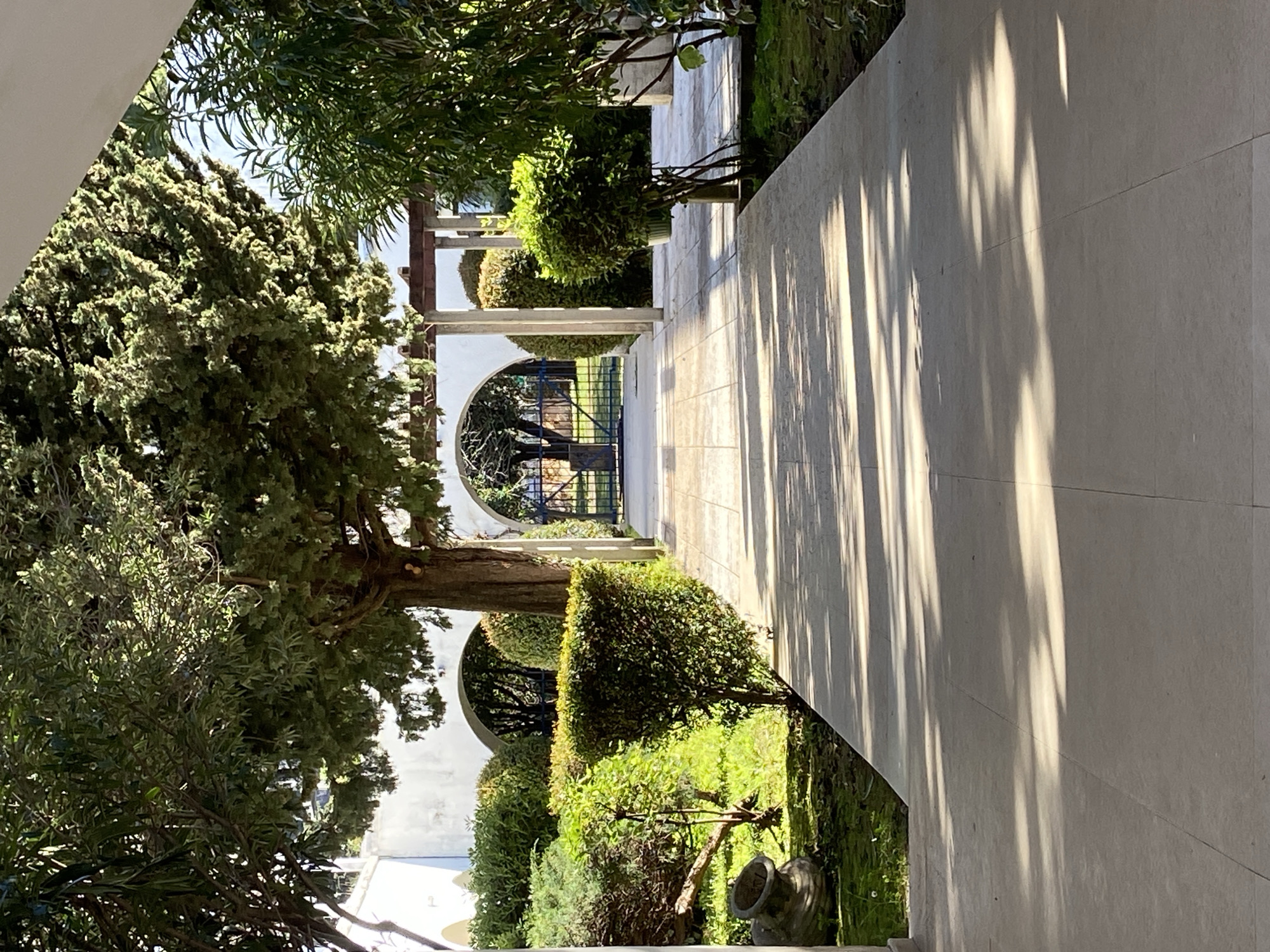 The image shows a garden pathway with white walls and arched gates in Budva, Montenegro. The pathway is made of light-colored stone slabs and is partially shaded by trees and foliage. The sun casts long shadows along the path. Manicured bushes and trees line the walkway, and there are views of the sea in the distance through the arched gates.