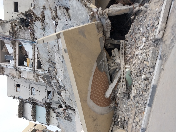 The image depicts a severely damaged building, likely due to an earthquake or bombing. The structure is reduced to rubble and debris, with floors collapsed and walls crumbled. A large, detached slab of concrete rests against the wreckage. The scene conveys a sense of devastation and destruction, highlighting the vulnerability of buildings in such circumstances.