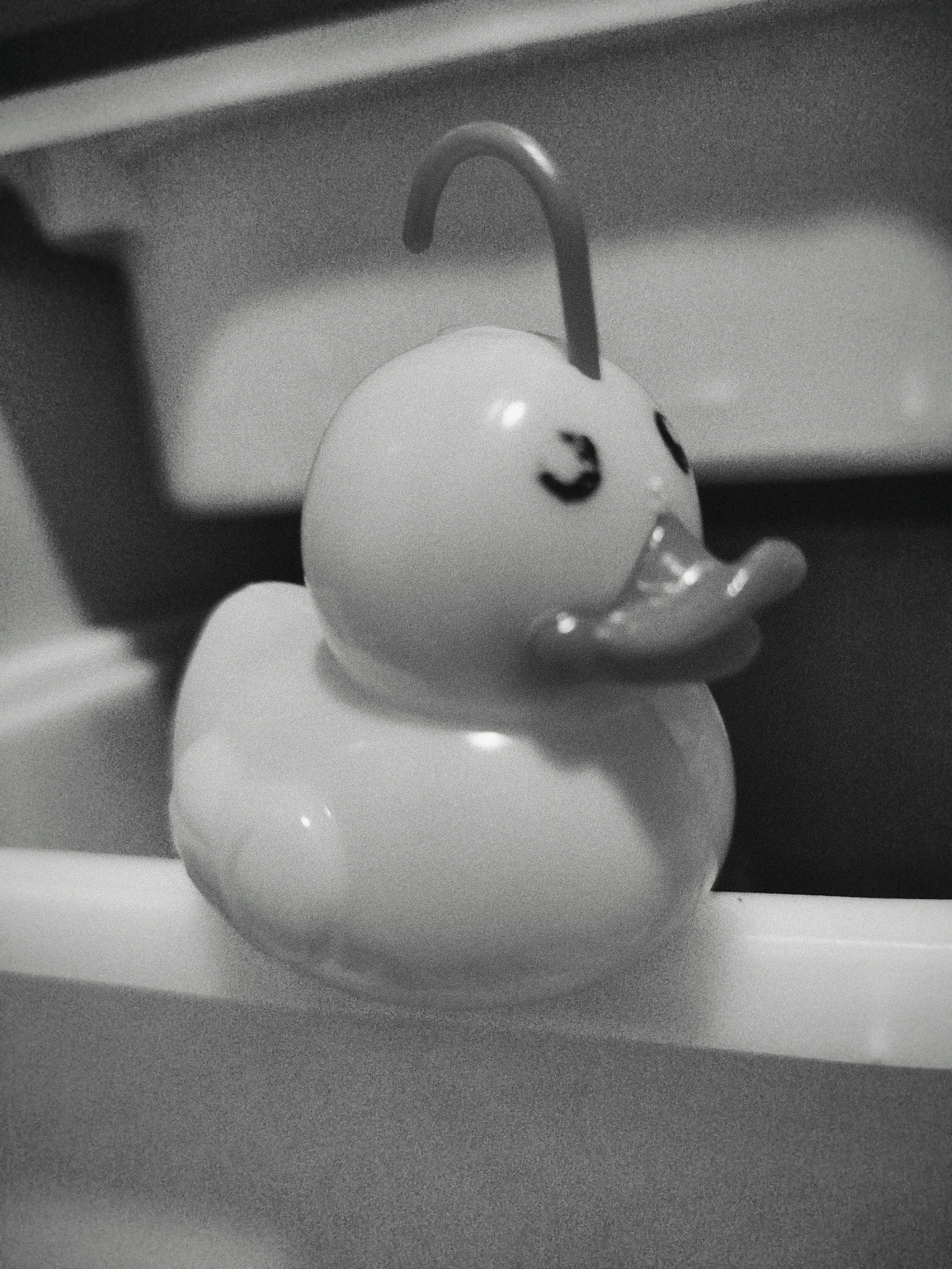 The image shows a close-up, black and white photograph of a yellow rubber duck toy. The duck is facing to the right, with its beak slightly open. It has two small, black eyes. A curved, orange handle extends upwards from the top of the duck's head. The duck is positioned on a white surface, possibly the edge of a bathtub or sink. The background is blurred and appears to be a textured, gray surface. The lighting creates a slight reflection on the duck's smooth surface. The overall impression is one of a simple, common object captured in a somewhat artistic, minimalist style. The image does not provide any information about its location in Mojoagung, Indonesia, or any people or actions.