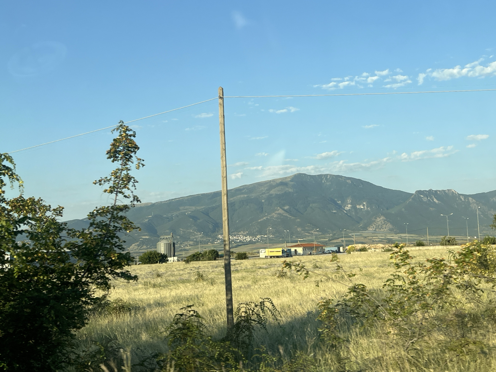 The image depicts a landscape near Prosotsani, Greece, under a clear blue sky with scattered white clouds. In the foreground, a tall wooden utility pole stands centrally, supporting a taut wire that stretches horizontally across the frame. To the left, the branches of a leafy green tree are visible, with some branches extending upwards and bearing small green and yellow leaves. The middle ground is dominated by a vast field of tall, dry, golden-brown grass, suggesting a rural or agricultural setting.
Further back, along a paved road, are several structures. A large, cylindrical industrial silo, likely for grain storage, is prominent on the left. Adjacent to it are buildings with orange roofs, one of which appears to be a small structure with a red-tiled roof and white walls. A yellow truck with a white cabin is parked near these buildings. Streetlights are visible along the road. In the background, a range of imposing, dark green mountains with rugged peaks rises against the horizon, with a small, white-housed village nestled at the base of the mountains. The overall scene suggests a sunny day in a semi-rural area with some industrial presence.
