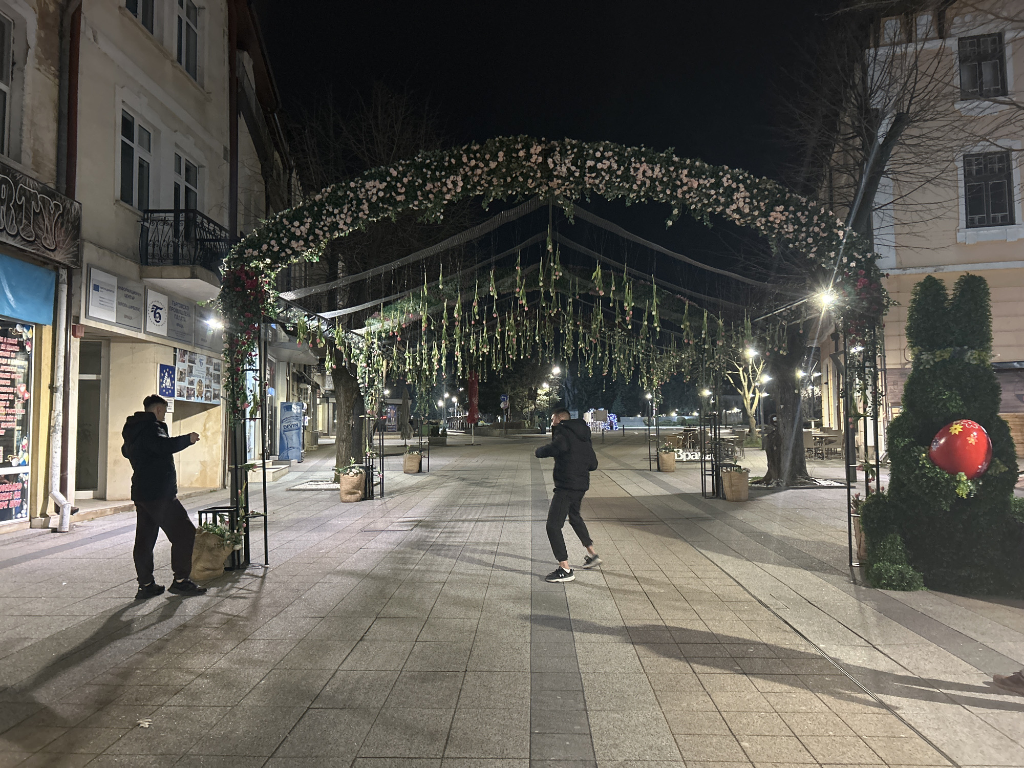 The image depicts an evening scene on a pedestrian walkway in Vratsa, Bulgaria. The walkway is lined with arched structures adorned with flowers and hanging decorations. Two men are present: one is standing near the entrance of the walkway on the left, while the other is positioned further down the walkway in a dynamic pose, possibly gesturing or moving. The environment includes shops and buildings on either side of the walkway, street lights, and decorative shrubbery, one with a large red ball attached. The walkway is paved with large square stones. The overall ambiance suggests a public space decorated for a festive occasion.
