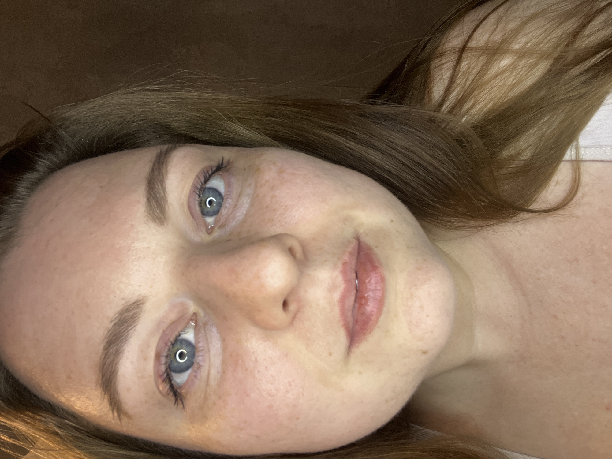 A close-up, angled portrait shows a light-skinned woman with light brown hair and blue eyes. Her face is freckled, and she has fair eyebrows and eyelashes with minimal mascara. The eyes reflect a circular light source. Her lips are pink and appear moisturized. The background is blurry and dark brown. The photo was not taken in Razdolye, Russia, as no geographic information is provided.