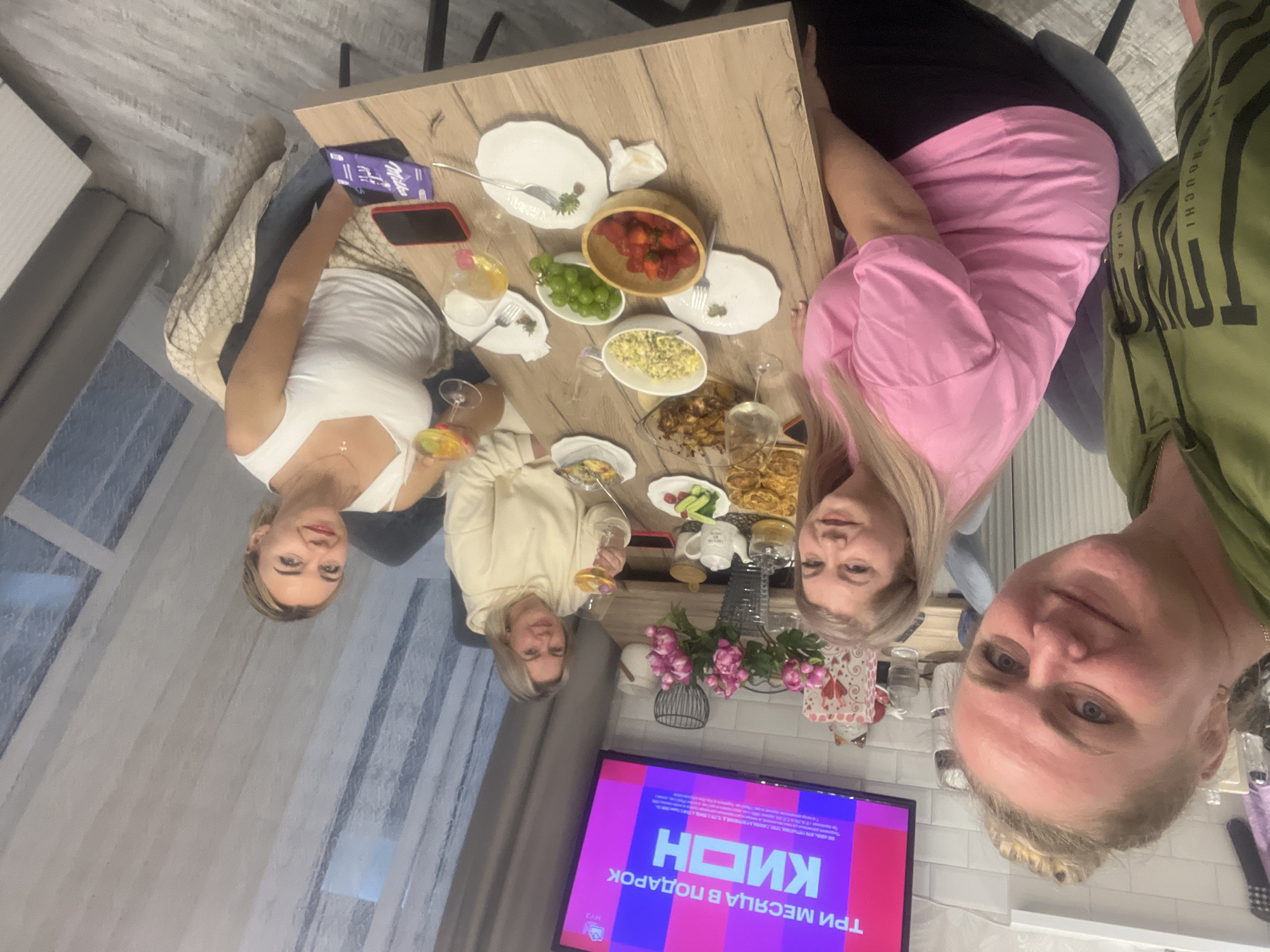 Four adult women are gathered around a wooden dining table in a domestic setting, located in Старые Кены, Russia. The woman on the far left, wearing a green hoodie with a black print, is taking a selfie of the group. She has blonde hair tied back and is smiling. To her right, a woman in a pink t-shirt and black pants, with blonde hair, is seated and smiling towards the camera. Further down the table, a woman in a light-colored hoodie holds a wine glass containing a yellowish liquid, looking towards the camera with a slight smile. On the far right, a woman in a white tank top and patterned trousers holds a cocktail glass with a colorful drink, also smiling towards the camera.
The rectangular wooden table is covered with various food items and tableware. These include a wooden bowl filled with strawberries, a white bowl with green grapes, a white bowl containing a salad (appearing to be corn/egg-based), a glass baking dish with roasted food, and a plate of fried items. Multiple white plates, forks, wine glasses, and cocktail glasses are distributed across the table. Also present are a white teapot with a matching lidded container, a small plate with sliced cucumbers and tomatoes, two smartphones (one with a red case, one black), a purple Milka chocolate bar, and white napkins. A vase containing pink artificial peonies is centrally placed on the table.
In the background, a flat-screen television mounted on the wall displays Russian text: "ТРИ МЕСЯЦА В ПОДАРОК" (THREE MONTHS AS A GIFT) and "КИОН" (KION), against a purple and pink graphic. White tiled walls are visible behind the table, extending to a kitchen counter area where decorative items, including a patterned box and a small figurine, are placed. Light-colored vertically striped curtains cover a window on the right side of the frame. The floor is light-toned, resembling wood planks or tiles. Grey dining chairs with black legs are positioned around the table. A white radiator or heating panel is partially visible on the left wall behind the selfie-taker.