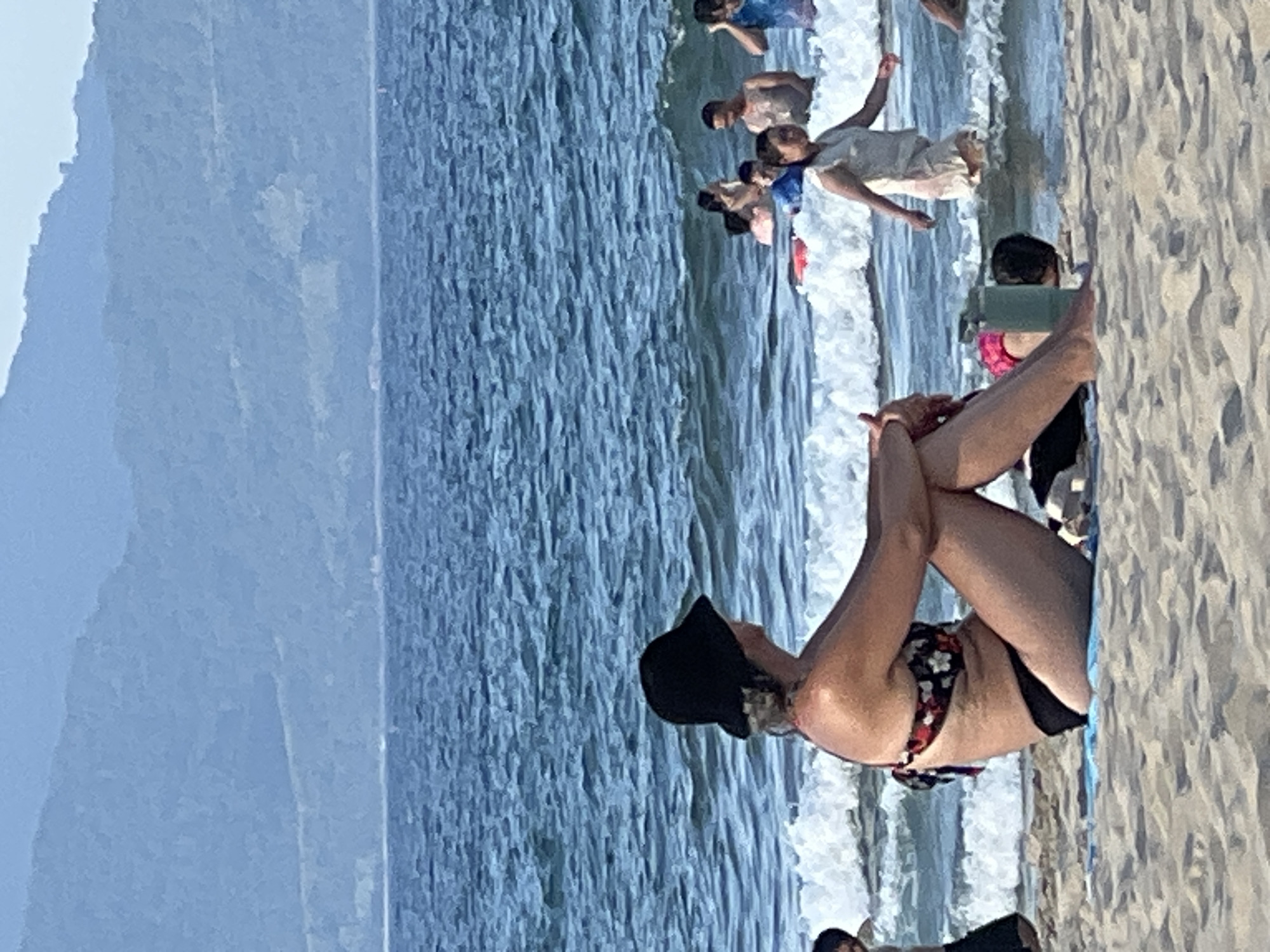 The image depicts a beach scene in Da Nang, Vietnam, with a woman in the foreground sitting on the sand and facing away from the camera. She is wearing a black bucket hat and a floral bikini top with black bottoms. The sea is in the background, with waves breaking on the shore. Several people are visible in the water, some swimming and others standing. In the distance, mountains form the horizon. The sand is light brown and appears to be wet near the water's edge. A light blue towel or mat is placed under the woman. A green water bottle is situated next to a person in the sand to her right. The overall atmosphere suggests a warm, sunny day at the beach.