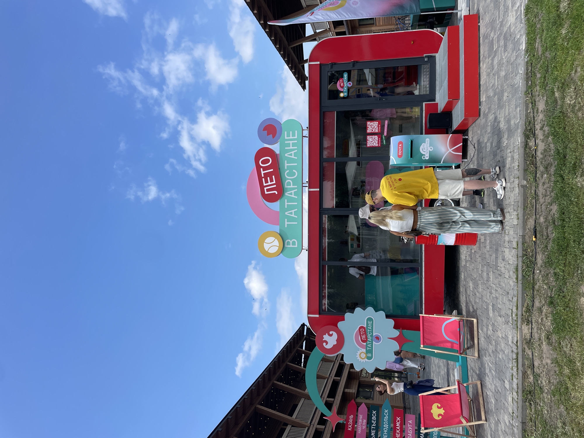 An outdoor scene features a red kiosk with the text "ЛЕТО В ТАТАРСТАНЕ" (Summer in Tatarstan) prominently displayed on its facade and roof. Two beach chairs, one red with a yellow symbol and another red with a blue design, are positioned in front of the kiosk. A young woman in a white top and blue pants stands to the left, taking a photo with her phone. A couple, a man in a yellow t-shirt and shorts and a woman in a white crop top and striped pants, are interacting near the kiosk's entrance. A blue mailbox is situated next to the kiosk. The background shows a modern wooden building with an arched structure and various directional signs. The sky is blue with scattered white clouds. The ground is paved with cobblestones and has patches of grass. The overall scene suggests a promotional event or attraction in Kazan, Russia, during the summer.