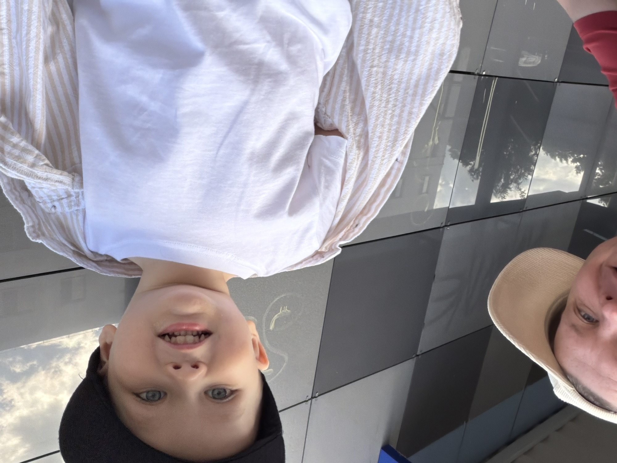A boy wearing a black baseball cap and a white t-shirt is captured in a close-up shot, his head tilted to the side. He has blue eyes and is smiling, showing his teeth. A striped shirt is draped over his shoulders. The background is a modern building facade with alternating dark grey and light grey rectangular panels. Another person, wearing a beige cap, is partially visible at the bottom of the image. The image appears to be taken outdoors in an urban setting. There is no specific information to determine the location in the city of Chaikovsky, Russia.