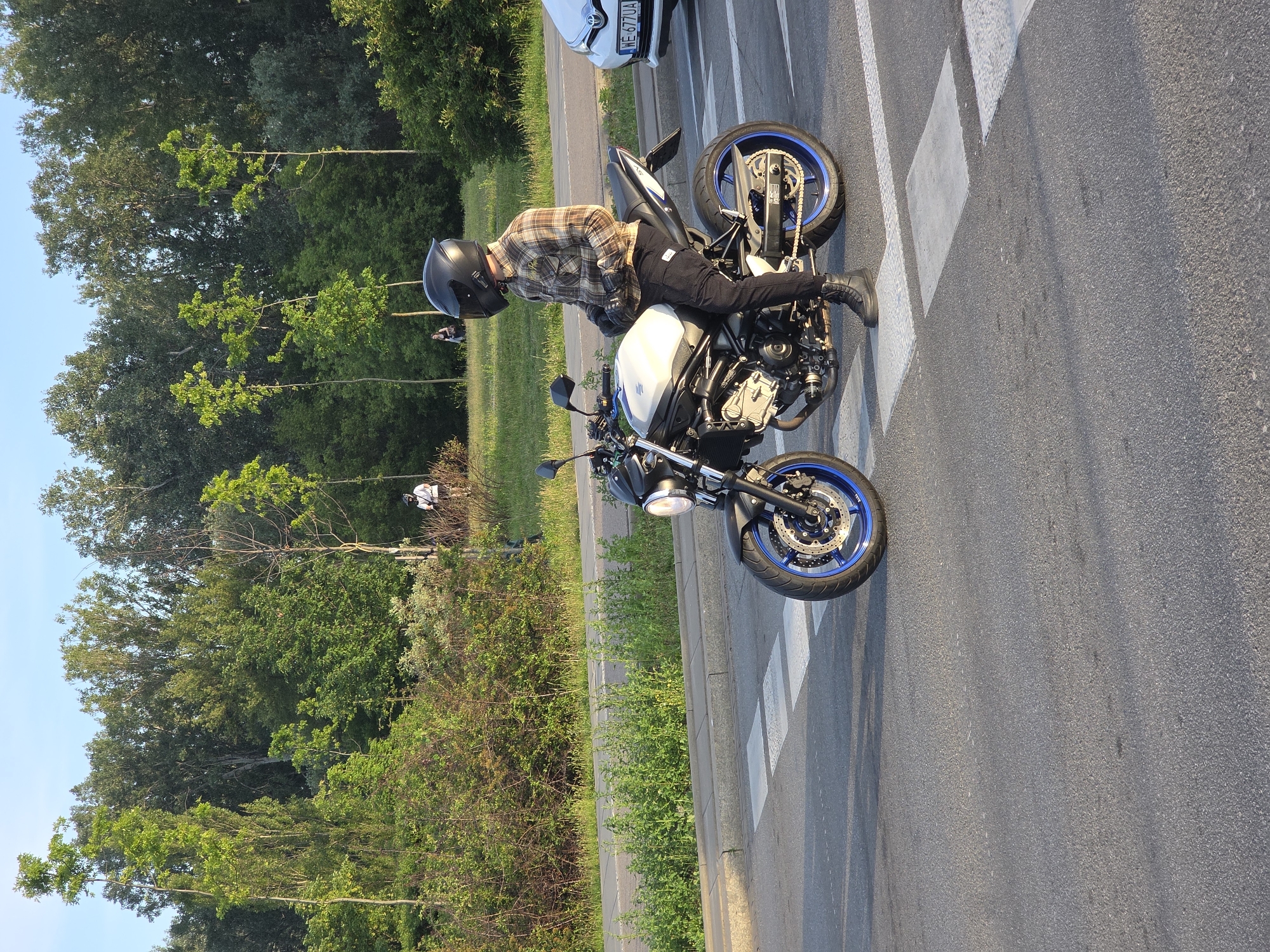 A person wearing a plaid shirt, black pants, black boots, and a black helmet sits on a white and black motorcycle. The motorcycle has blue rims. The person is facing away from the camera, looking to the right. The motorcycle is parked on an asphalt road, with white pedestrian crossing lines on the road. To the left of the road is a grassy area with trees and bushes. In the background, a white car is visible. The scene appears to be outdoors during the daytime, possibly late afternoon or early evening given the lighting. The weather seems pleasant and sunny. The image is oriented vertically. The text "WE 677UA" is visible on the license plate of the white car.