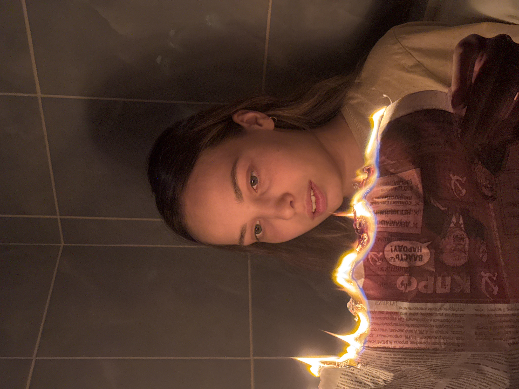 In a dimly lit bathroom with grey tiled walls, a young woman with long, brown hair holds a burning newspaper. The flames, intensely bright and casting an orange glow, consume the top edge of the newspaper, revealing Russian text and images of individuals. Her gaze is directed downwards at the burning paper, her mouth slightly open in an expression of curiosity or concern. The woman is wearing a tan t-shirt, and a small hoop earring is visible in her left ear. The overall scene suggests a moment of personal reflection or symbolic act within a domestic setting in Chaikovsky, Russia.