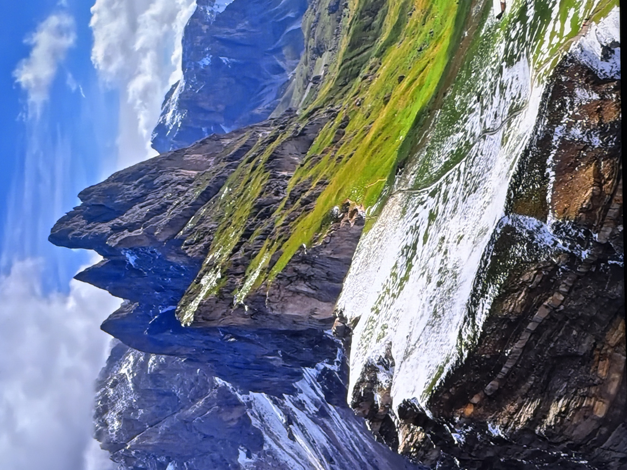 The image depicts a dramatic mountain landscape with jagged peaks, steep slopes, and patches of snow. The mountains are layered with various rock formations, and some slopes are covered in vibrant green grass. The sky is blue with scattered white clouds. There are no visible people, structures, or signs of human activity within the immediate mountain scene. The scene is entirely natural and does not contain any elements that would place it within the city of Chirchik, Uzbekistan.