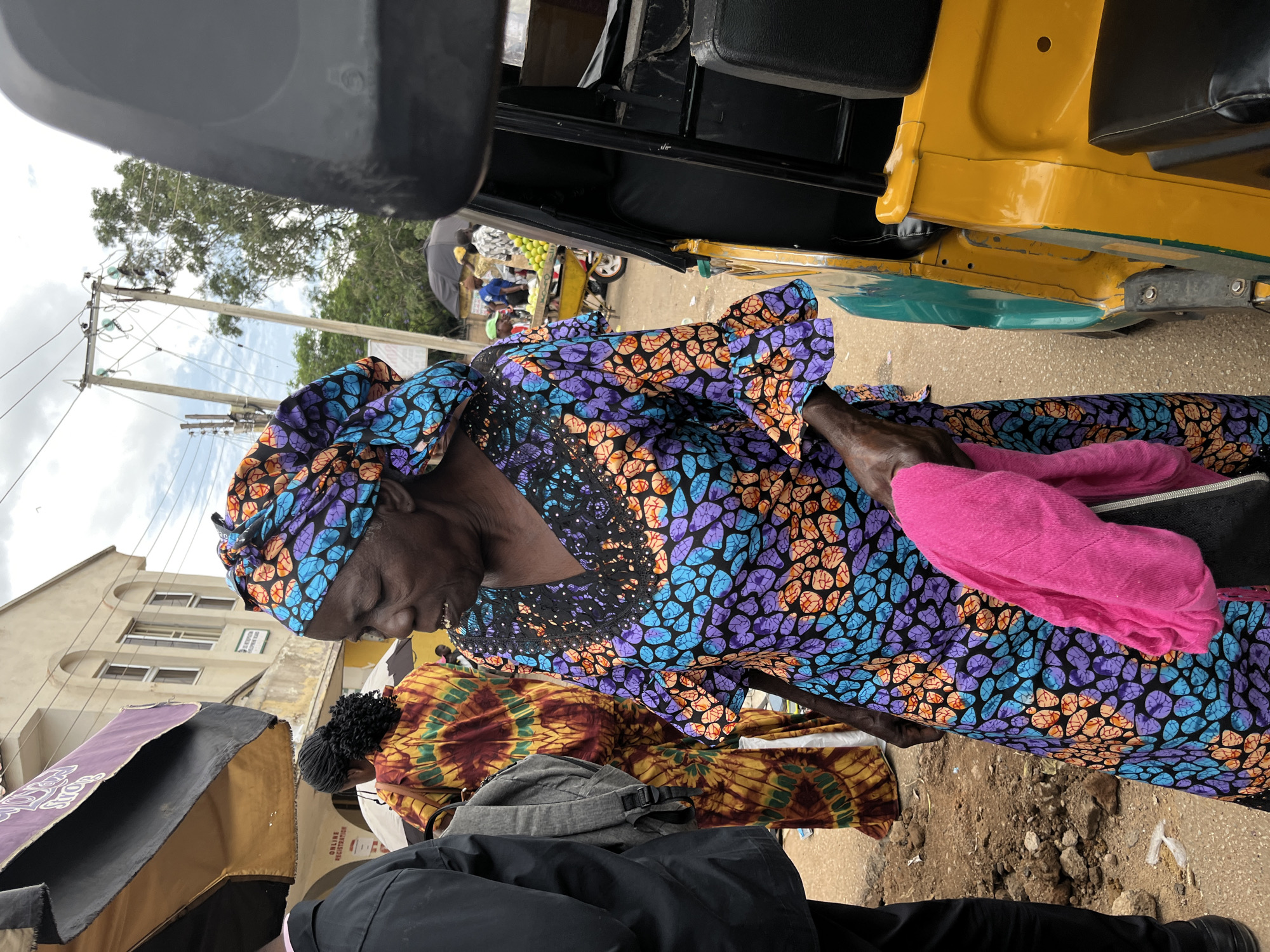 This detailed description pertains to a street scene in the city of Jos, Nigeria.
The image prominently features an older Black woman positioned in the foreground, slightly left of center, facing partially towards the viewer's left. She wears a vibrant, long-sleeved dress with a V-neck and ruffled cuffs, patterned in a mosaic-like design of black, blue, purple, and orange amorphous shapes. A matching headwrap covers her hair. Small white flecks are visible around her mouth. She holds a folded bright pink textile in her left hand, which also grips a small black bag or pouch.
To the immediate left of the main subject, a second individual, largely obscured, wears a black long-sleeved shirt, black trousers, and black shoes. A grey backpack is slung over their left shoulder. Behind this person, a third individual, seen from the back, wears a loose-fitting garment with a tie-dye pattern in yellow, red, and brown hues.
To the right of the main subject, the front portion of a yellow and teal/green three-wheeled auto-rickshaw (locally known as a Keke Napep) is visible, including a black vinyl seat and the side body panel.
In the mid-ground, under a black umbrella, several blurred figures are present. A yellow wheelbarrow filled with green spherical fruits (possibly limes or similar citrus) is visible, with a person in a green cap standing nearby. Overhead, power lines are strung between two utility poles against a cloudy sky. The background includes a light-colored, two-story building with arched windows and a partially visible sign that reads "...METROPOLITAN..." The ground is uneven, a mixture of paved areas and bare earth with scattered rocks and debris. Various shadows indicate overhead cover from structures or market umbrellas, one of which is black and yellow.