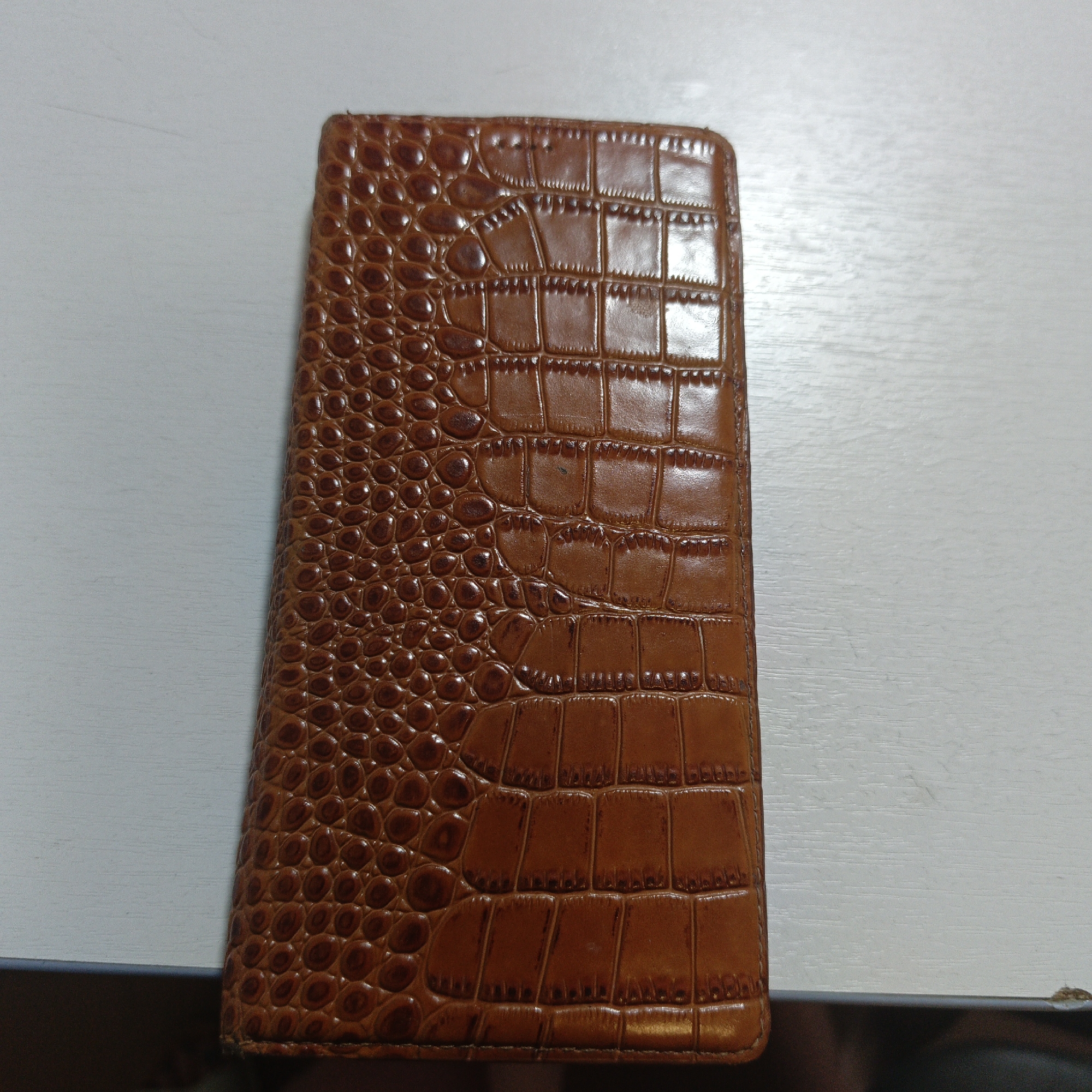 An overhead, close-up shot shows a brown, alligator-embossed leather wallet lying on a white, textured surface. The wallet is oriented vertically and fills the left side of the frame. The texture of the leather is clearly visible, with a pattern of raised scales and rectangles. The white surface appears to be a table or desk with a slight wood grain. There are no people or visible actions occurring in the image. The lighting is neutral, casting minimal shadows. No specific location within Ulan-Ude, Russia, can be determined from the image.