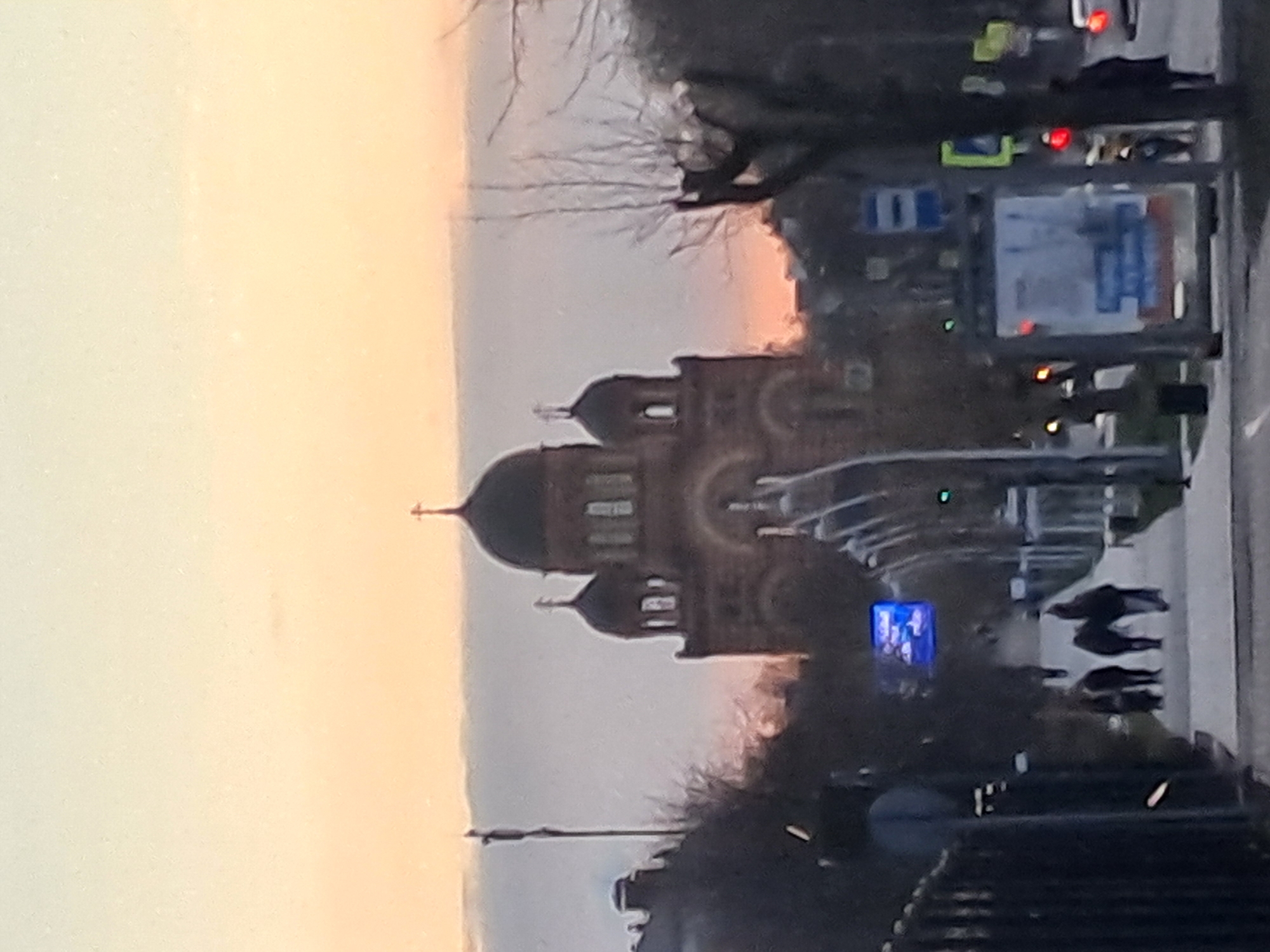 The image shows a blurry street scene in Elektrostal, Russia. An Orthodox church with onion domes is visible in the background against a hazy sky. Street lights, traffic signals, and road signs are present along the right side of the image. People are walking on the sidewalk. Vehicles are also visible on the road.