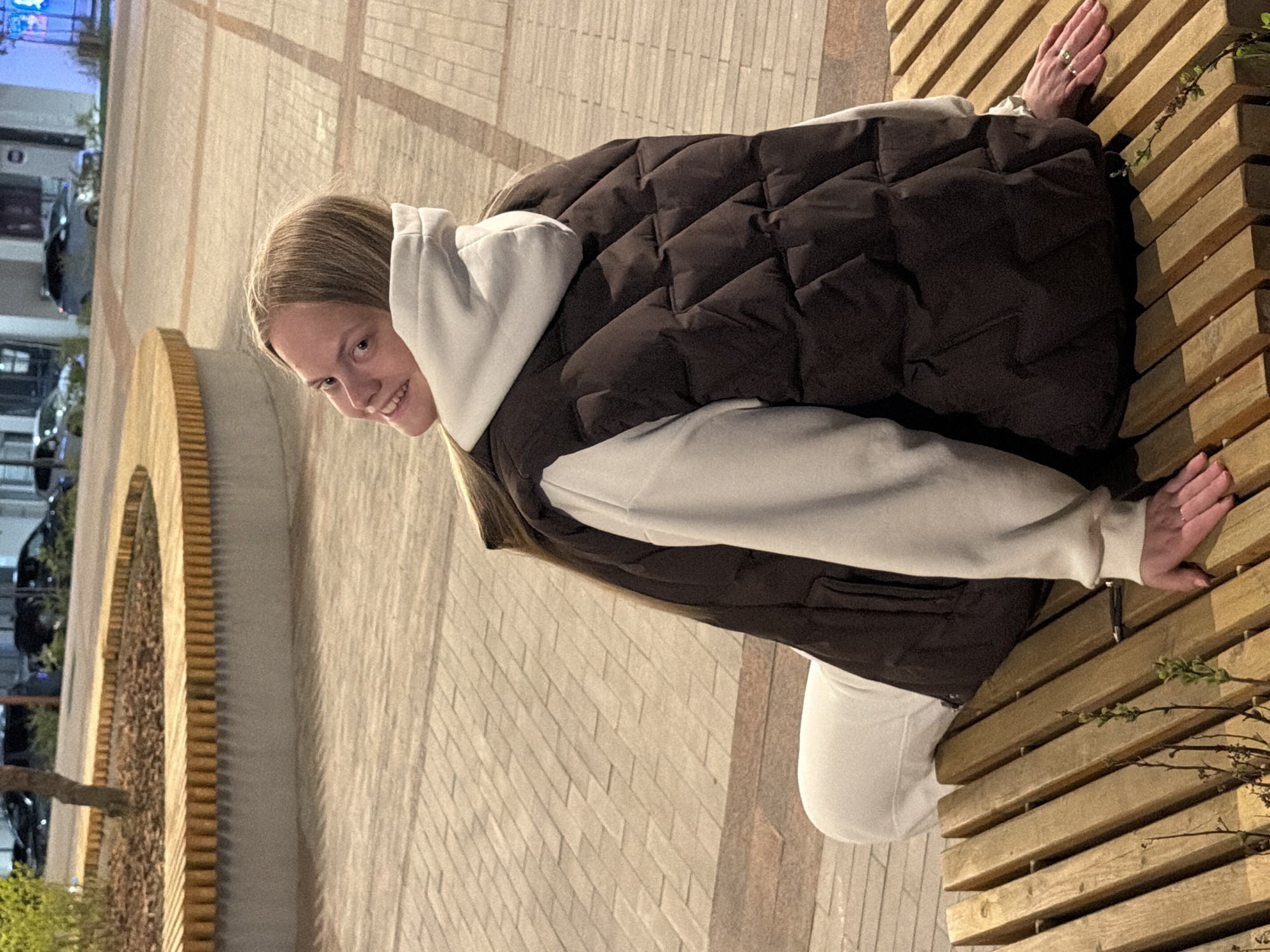 A young woman in Izhevsk, Russia, is posing on a wooden bench in an urban setting. She is wearing a white hooded sweatshirt and a dark brown quilted vest, and white pants. Her left hand rests on the bench behind her, and her right hand is placed on the front of the bench. A paved walkway is next to the bench. A distinctive semicircular wall with an ornate pattern is visible in the background. Cars are parked in the distance under streetlights.