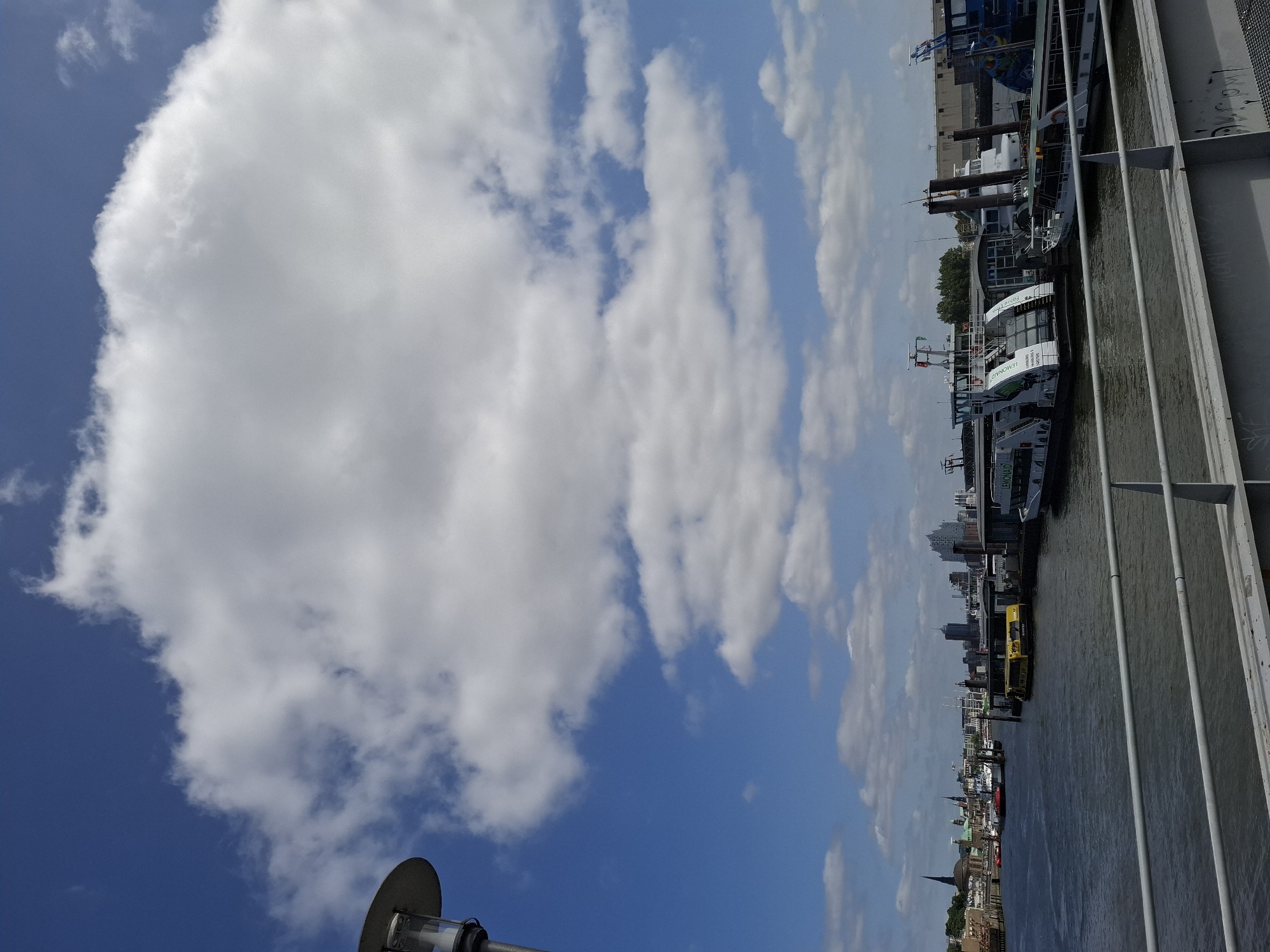This is a landscape image captured on a sunny day, likely in Hamburg, Germany, given the prominent Elbphilharmonie concert hall visible in the background. The scene depicts a wide river, possibly the Elbe, with several boats and ferries docked along the waterfront.
In the foreground, a modern metal railing runs across the bottom of the frame, suggesting the vantage point is from a bridge or pier. The water is a deep blue, reflecting the bright sky above.
Further out in the river, a yellow ferry with visible markings is docked. To its right, a larger white ferry, labeled "LEMONAID," is also moored. Several other boats of various sizes are present in the middle ground.
The background showcases the Hamburg skyline, characterized by a mix of architectural styles. The distinctive silhouette of the Elbphilharmonie concert hall stands out with its glass facade. Other buildings, including church spires and modern skyscrapers, contribute to the urban panorama.
The sky is a vibrant blue, partially obscured by large, fluffy white clouds. A lamppost with a modern design is visible on the far left of the image, its metallic head peeking into the frame. The overall atmosphere is one of a bustling port city on a clear, pleasant day. There are no visible people in the image.