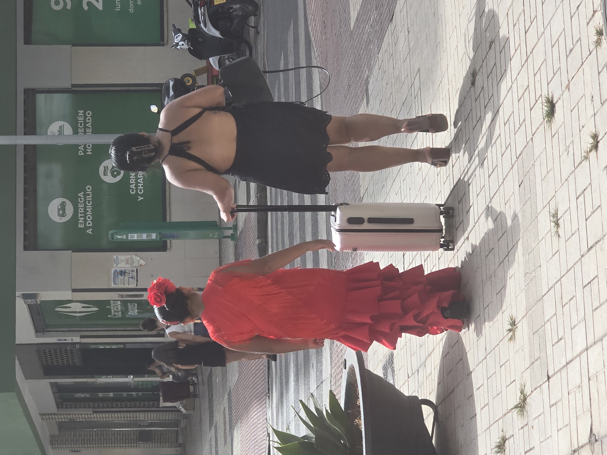 Two women walk along a paved sidewalk in Málaga, Spain. The woman on the left wears a vibrant red, ruffled flamenco-style dress and has a large red flower in her hair. She is pulling a small, white rolling suitcase with her left hand. The woman on the right wears a black, open-back dress and carries a black handbag over her left shoulder. She is also pulling a small, white rolling suitcase with her right hand. They are walking away from the camera, on a sidewalk in front of a building with green signage that includes the Carrefour Express logo and advertisements for home delivery services. A plant in a large dark pot is visible to the left of the women. In the background, scooters are parked and other pedestrians are visible in the distance. The scene appears to be a sunny day.