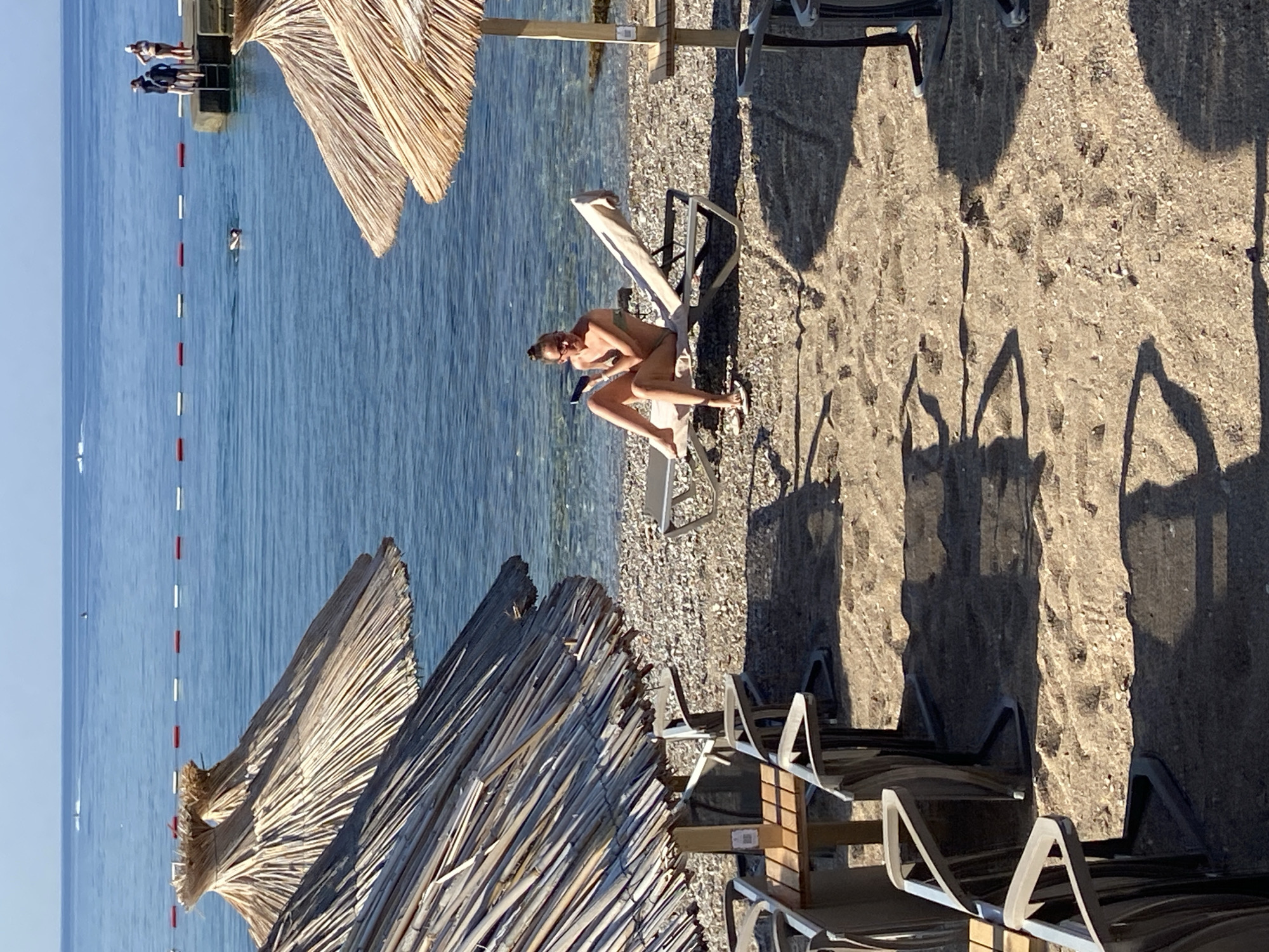 A woman is relaxing on a sun lounger on a beach in Budva, Montenegro. She is wearing a bikini and sunglasses and appears to be reading a book. The beach is made of pebbles and there are several other sun loungers and umbrellas visible. The sea is calm and blue, and the sky is clear. In the distance, there is a pier with a few people standing on it. The time of day is likely late morning or early afternoon, as indicated by the strong shadows cast by the umbrellas and loungers. The weather appears to be warm and sunny, perfect for a day at the beach. The overall mood is one of relaxation and enjoyment. There is no visible text in the image.