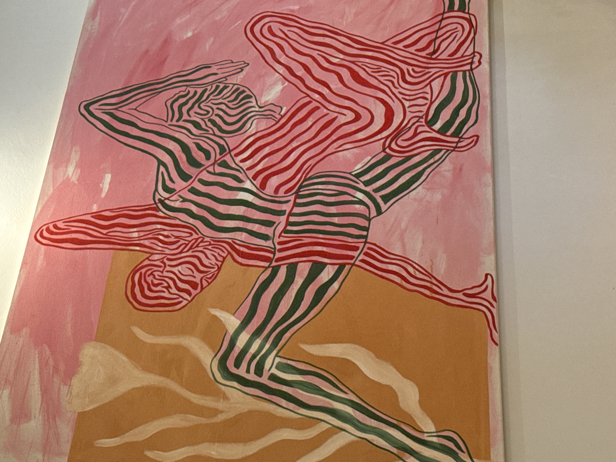An abstract painting, oriented horizontally, is mounted on a plain white wall, illuminated by overhead lighting. The artwork features two stylized human figures rendered with bold, irregular stripes of varying colors. The figure on the left is predominantly striped in green and pink, depicted with a bent leg and arm extended upwards. The figure on the right is primarily striped in red and pink, appearing to interact or intertwine with the first, resting on its side with limbs curled. The background of the painting is divided into two distinct color fields: an ochre-toned left section displaying light beige, organic, plant-like forms, and a soft pink right section. This scene is located in the city of London, United Kingdom.