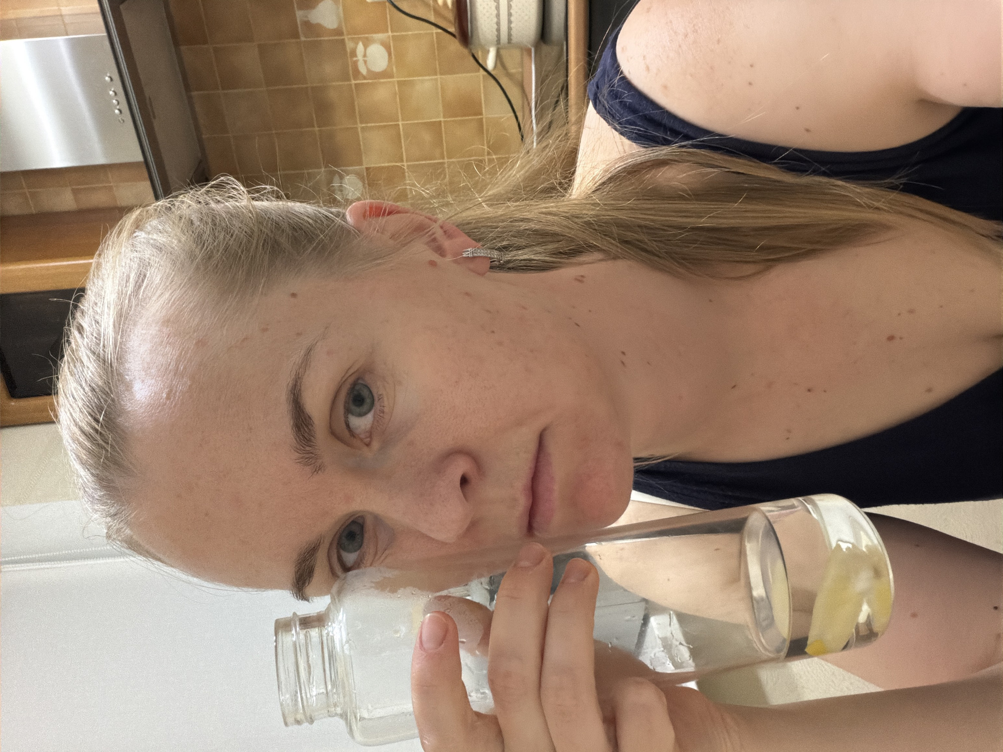A young woman with blonde hair tied back in a ponytail, wearing a dark blue top and hoop earrings, is holding a clear glass bottle filled with water and lemon slices. Her eyes are open, and she has a neutral expression. The background appears to be a kitchen, with tiled walls and a stainless steel range hood visible. There is no information in the image to suggest the location is Khabarovsk, Russia.