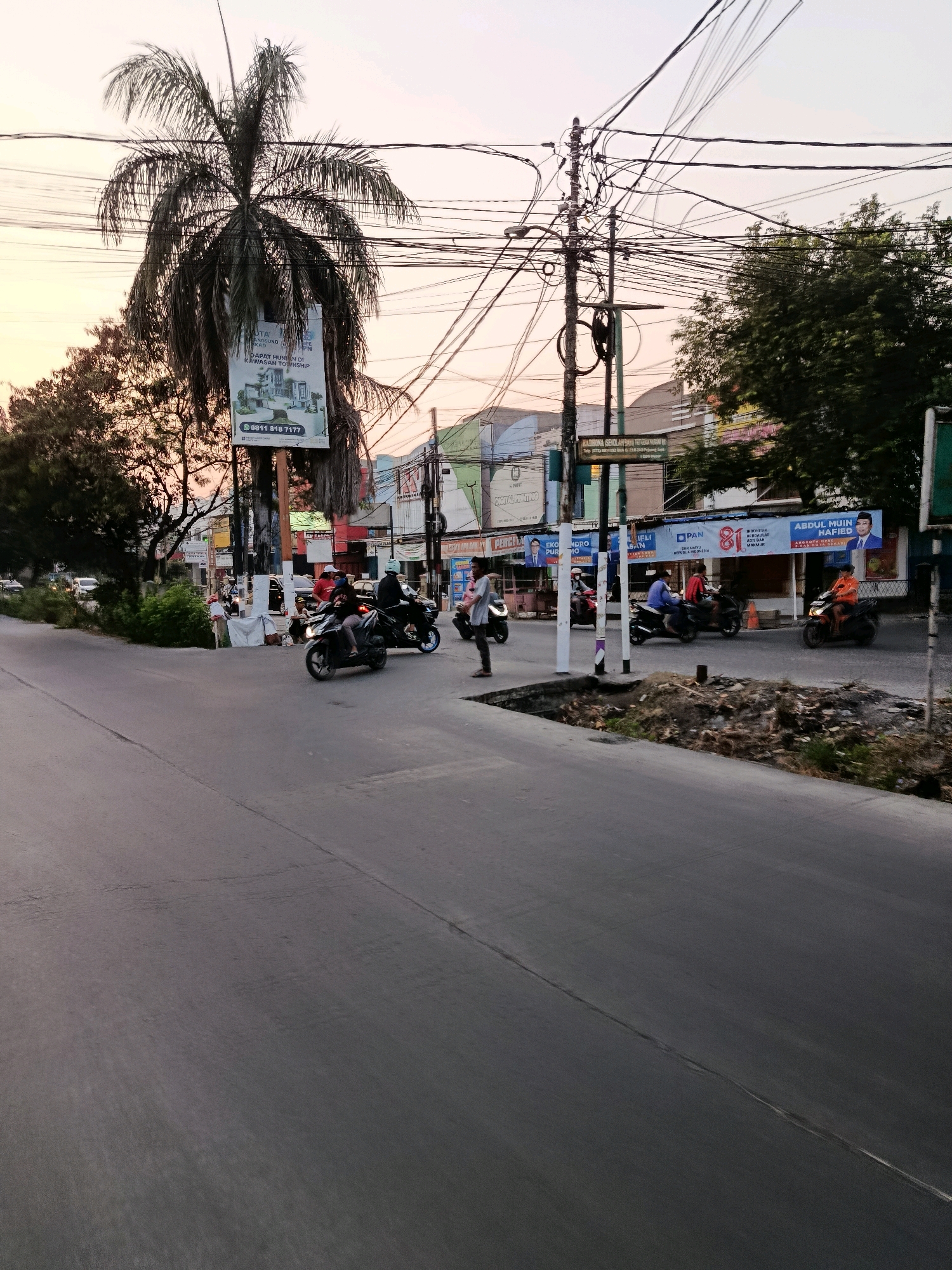 The image depicts a street scene in Bekasi, Indonesia, during what appears to be late afternoon or early evening. The sky is a pale orange and pink, indicating the setting sun. A prominent palm tree stands on the left side of the frame, with a large billboard attached to its trunk advertising real estate. The billboard features an image of a modern house and a phone number.
The street is paved with asphalt, and there is a distinct line dividing the lanes. Several people are present, mostly on or near motorcycles. On the right side of the road, a man in an orange shirt is riding a motorcycle. Further down the street, there are more motorcycles and pedestrians, with some individuals gathered near a small stand. Buildings line the street, displaying various signs and banners, some of which appear to be political advertisements. One banner prominently features a picture of a man with the text "ABDUL MUIN HAFID" and "ANGGOTA DPR RI PARTAI GERINDRA". Another banner is for "PAN". Electrical wires are strung across the sky, supported by utility poles, one of which is heavily laden with cables and appears to be a central point for the network. Trees with dark green foliage are visible behind the buildings and on the left side of the street. The overall environment suggests a busy urban street with commercial activity and residential areas nearby.