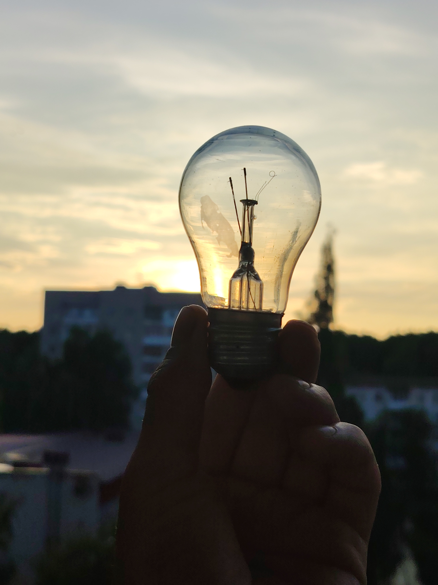 A close-up, silhouette shot captures a hand holding a clear glass light bulb against a dusky sky, with the sun setting behind a building in the background. The light bulb, with its filament visible, is the focal point, and it appears to be partially lit by the sunset. The background consists of an apartment building and trees, typical of a residential area. The image is likely taken from a window or balcony in Chaikovsky, Russia, given the architectural style of the building.