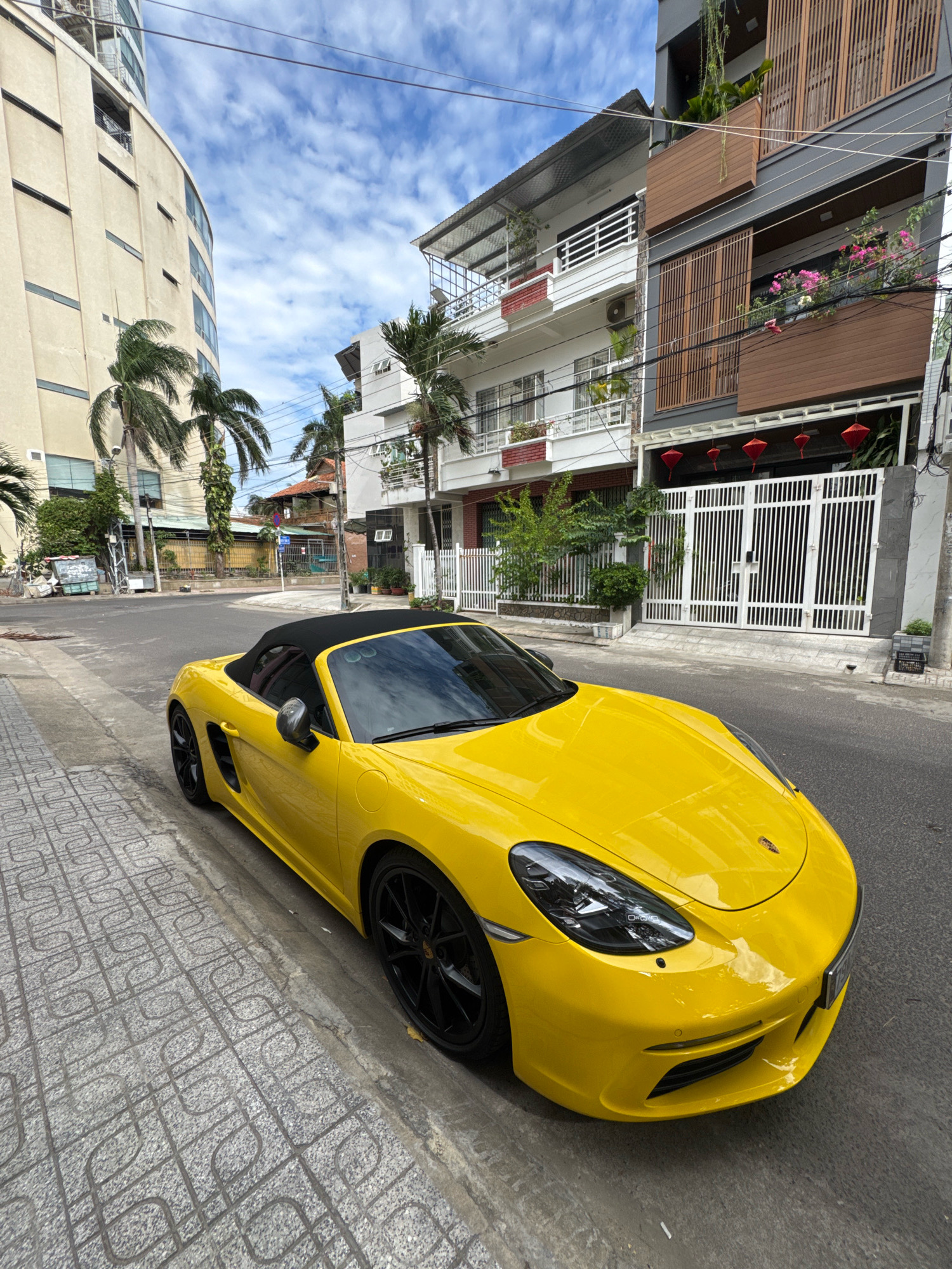 A bright yellow Porsche Boxster convertible is parked on a street in Nha Trang, Vietnam. The car is positioned on the left side of the frame, its front facing towards the right. The street has a gray, patterned sidewalk on the left and asphalt pavement on the right. The background shows several buildings, including a tall, curved cream-colored building on the far left and narrower, multi-story buildings with balconies and decorative wooden elements. Palm trees are visible between the buildings. The sky is blue with scattered white clouds. Red lanterns are hanging from the facade of one of the buildings. There are no people visible in the image.