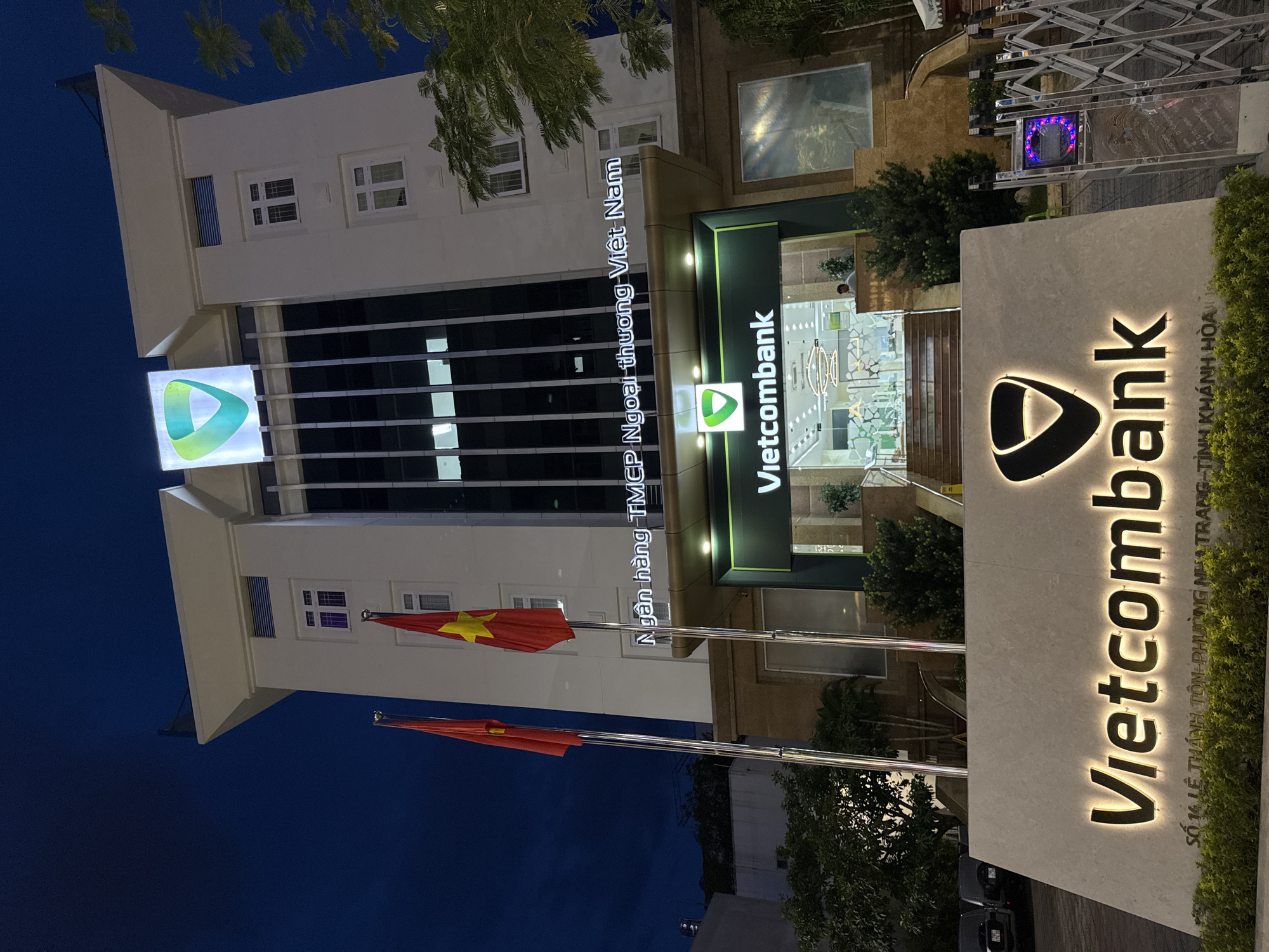 The image shows the Vietcombank building in Nha Trang, Vietnam, at night. The building is illuminated, and its facade features the Vietcombank logo. In front of the building is a large sign with the Vietcombank logo and the bank's name in illuminated lettering. The address "SO 14 LE THANH TON - PHUONG NHA TRANG - TINH KHANH HOA" is visible below the bank's name on the sign. Two Vietnamese flags are planted in front of the building. Trees and landscaping surround the area. Several cars are parked to the left of the frame. A security gate is visible to the right. The sky is dark, indicating it is nighttime.