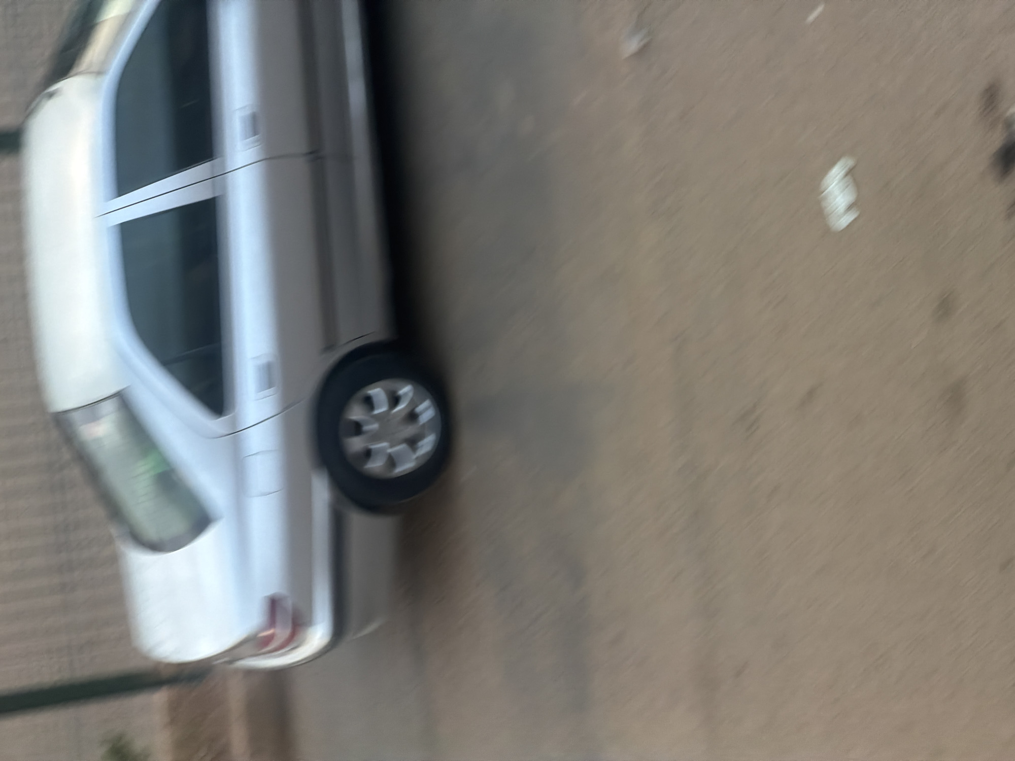 The image shows the rear passenger side of a silver sedan, likely a Peugeot 406, parked on a dirt road in Bukuru, Nigeria. The car is silver with tinted windows. The rear wheel is visible, featuring a silver multi-spoke alloy rim. In the background, a beige wall with vertical grooves is visible, possibly part of a building or fence. The ground is a light brown, dusty surface. There is a small white object on the ground near the bottom right of the frame, possibly a piece of trash. The image appears to be taken with a motion blur, suggesting the vehicle or the camera was moving. There are no people or other vehicles clearly visible in the frame.