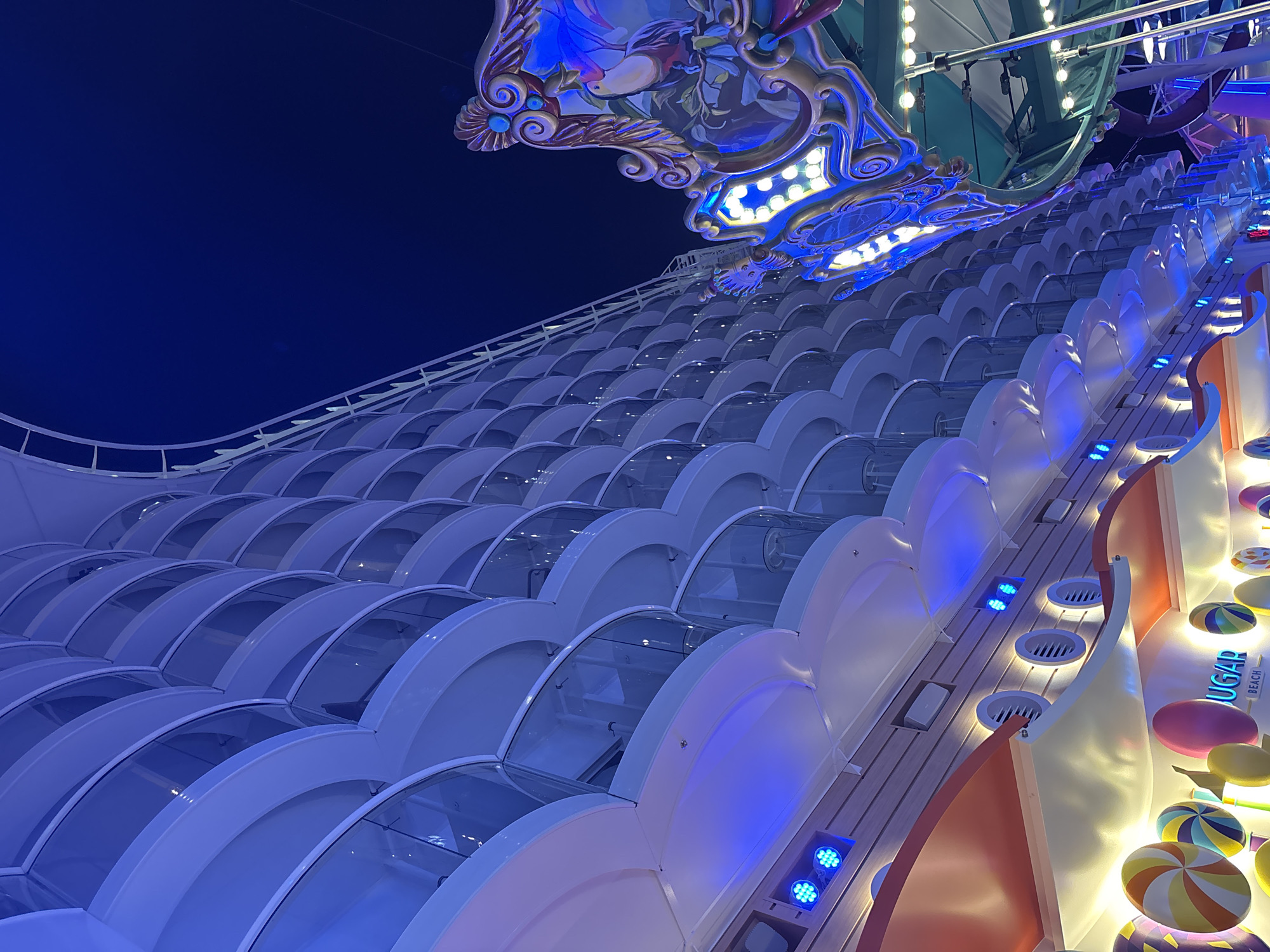 The image depicts a structure composed of a series of arched segments, possibly glass or a transparent material, arranged in a stepped, cascading manner. The architecture suggests a modern, possibly futuristic design. The lighting is predominantly blue, accentuating the curves and creating a visually striking pattern of light and shadow. In the foreground, a sign is partially visible, with the word "SUGAR" and "BEACH" discernible, suggesting this might be part of a sweet shop or a beach-themed attraction. Decorative elements, including colorful, oversized candy shapes, further reinforce this theme. The architectural style, the use of blue lighting, and the candy-themed decor suggest that this location might be part of a tourist attraction or entertainment complex in an urban setting.