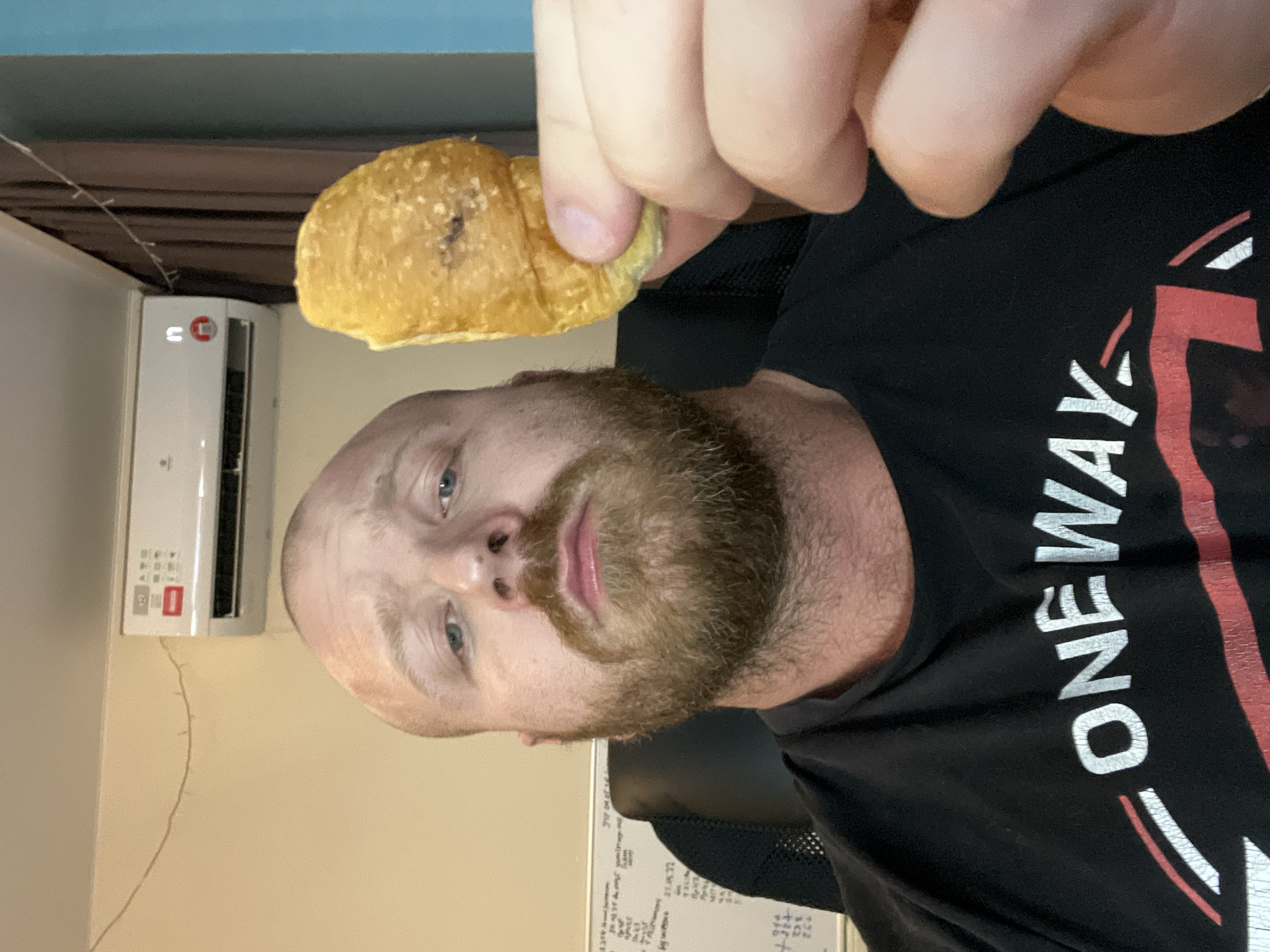 A man with a bald head and a beard is holding a croissant in his hand. He is wearing a black t-shirt with the word "ONE WAY" printed on it in white and red. Behind him is an air conditioner on a wall. There is a whiteboard in the background. The location is Noviy, Russia.