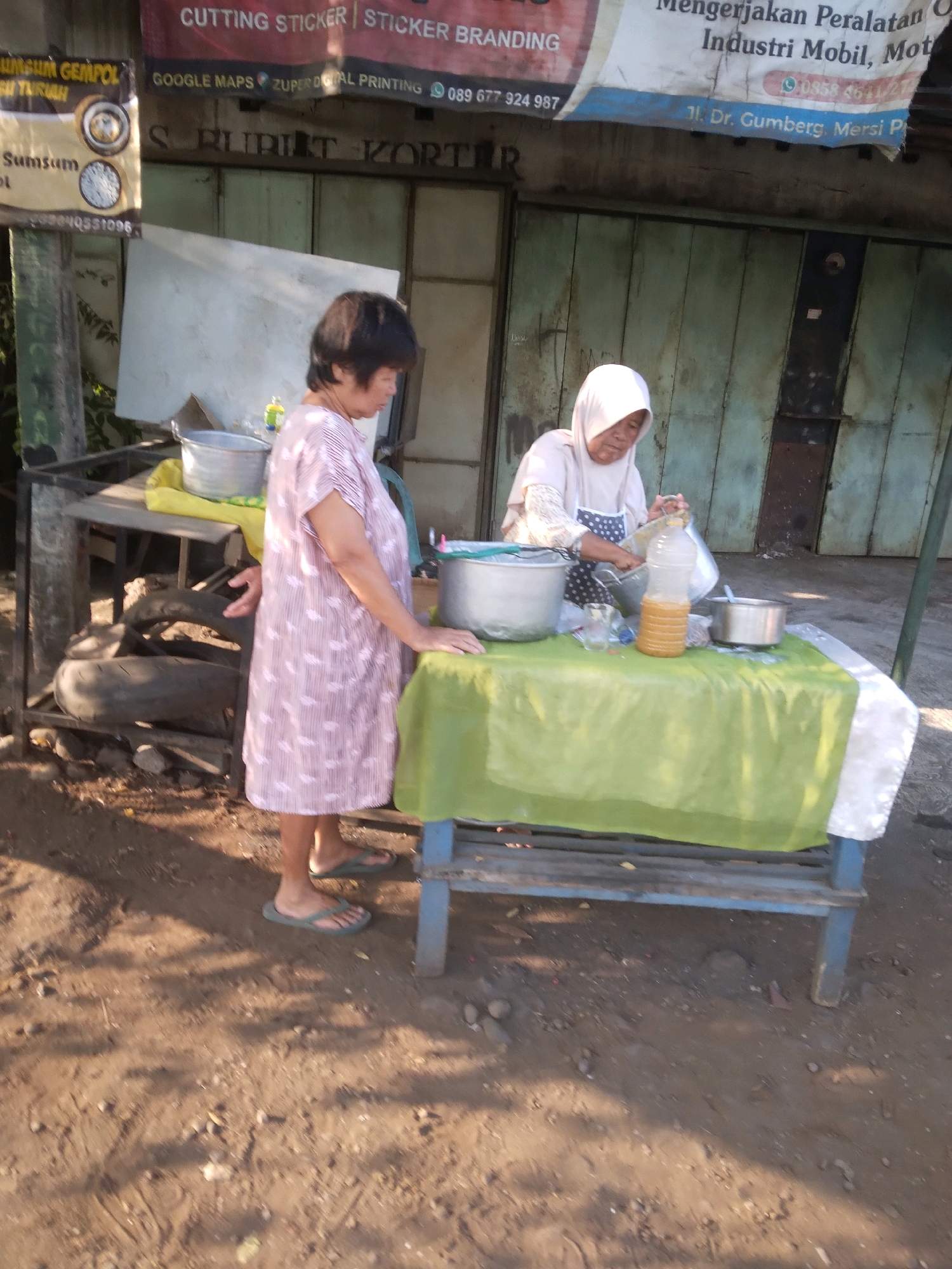 This image depicts an outdoor scene in Desa Pamijen Kecamatan Sokaraja, Indonesia. In the foreground, a woman in a pink and white patterned dress stands facing right, wearing green flip-flops. She is near a table covered with a green plastic sheet, on which several metal bowls, a plastic bottle filled with a yellow liquid, and a white object are placed. Behind her, to the left, is a metal rack with what appear to be stacked tires and other items. In the background, another woman wearing a white hijab and a white and blue patterned apron is seen from the chest up, leaning over the table and handling a metal bowl and the yellow liquid bottle. A partially visible banner hangs above the scene, displaying text in Indonesian. The ground is composed of dirt and scattered small stones. The overall environment suggests a market or roadside stall setting.