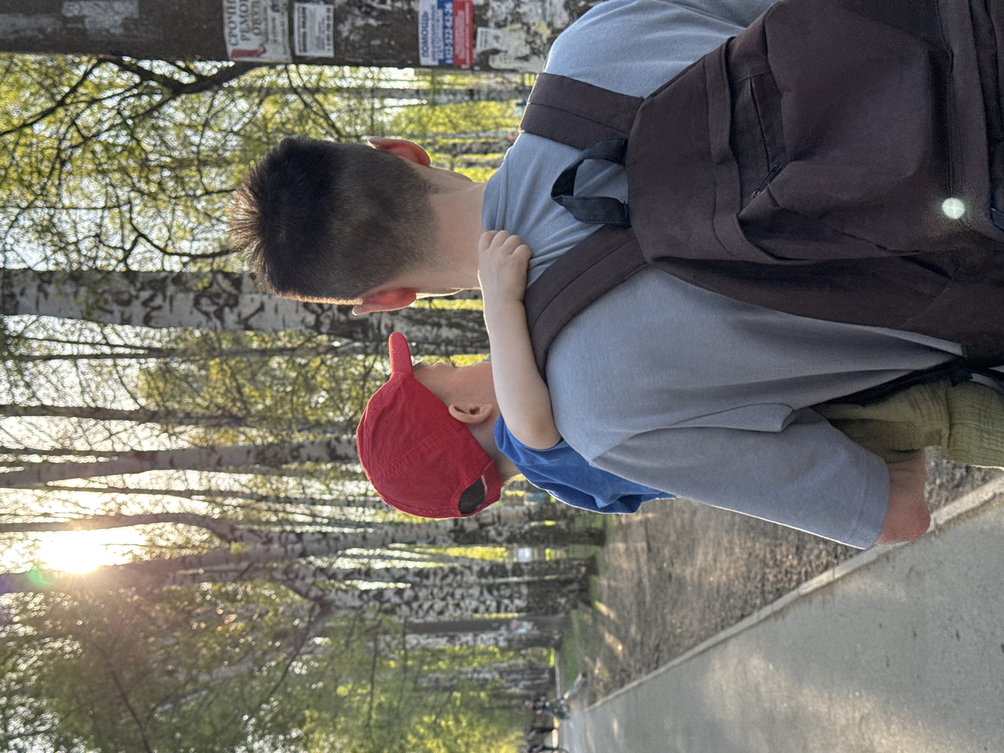 A person with a backpack is carrying a small child in the image. The child is wearing a red baseball cap. The location appears to be a park or wooded area with trees lining a pathway. The city may be Izhevsk, Russia, based on external knowledge. Sunlight is visible through the trees. The child is holding onto the person.