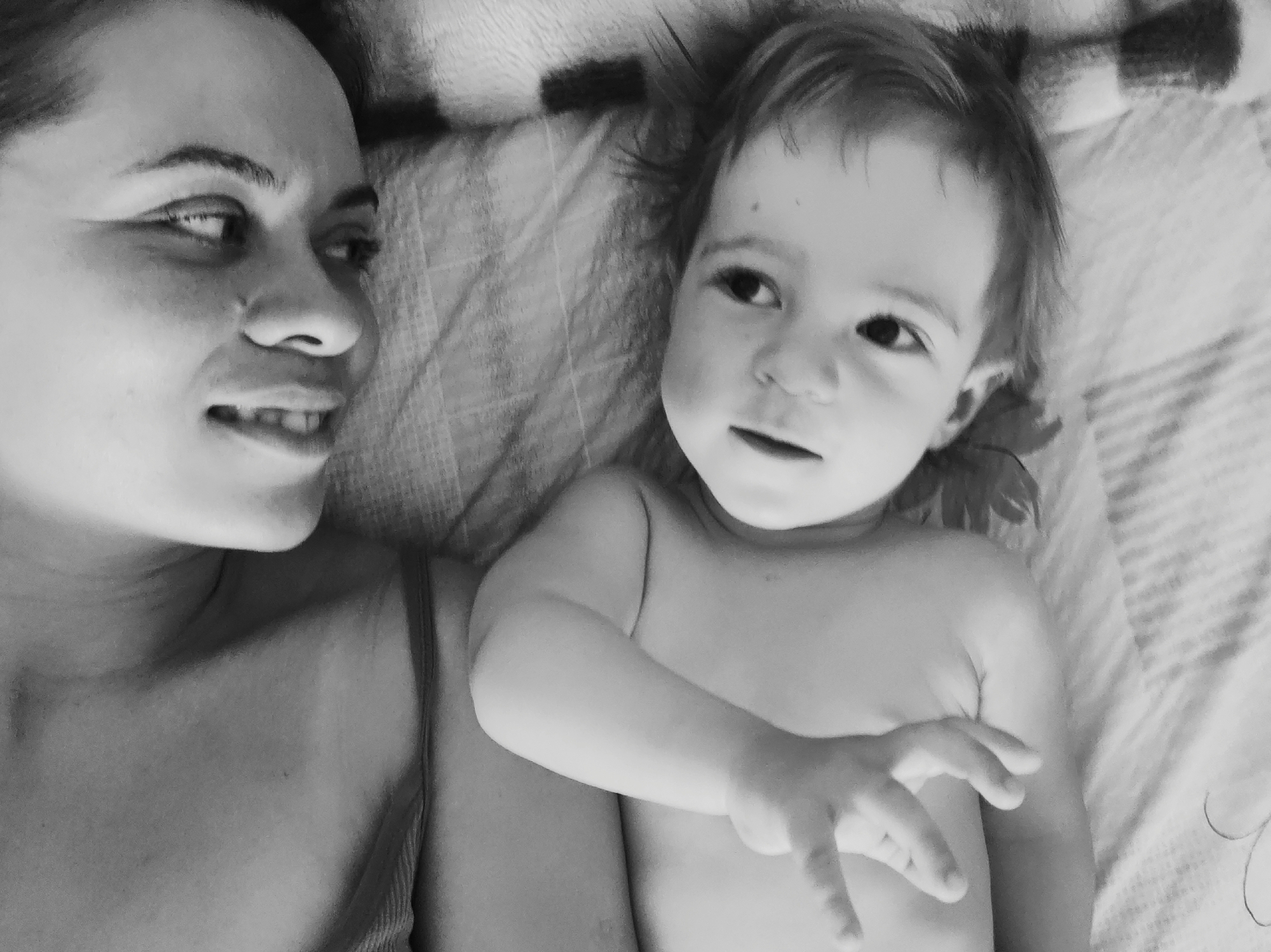 A black-and-white photograph depicts a woman and a young child lying on a patterned surface, likely a bed. The woman is positioned on the left side of the frame, facing the child with a partial smile. Her features are clearly visible, and her head is close to the edge of the image. The child is lying next to her, slightly angled towards the viewer. The child is partially unclothed, with one arm extended towards the camera. The background is a patterned surface, possibly a blanket or sheet. The location is Unknown, Unknown. The image captures a moment of interaction between the woman and the child.