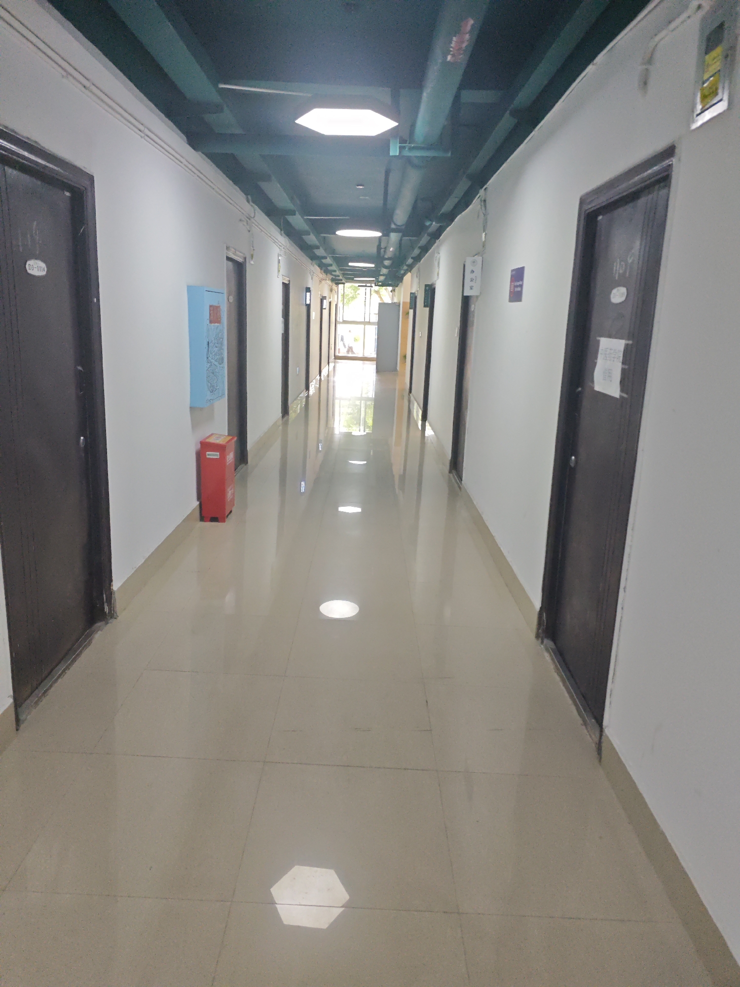 The image depicts a long, narrow, and brightly lit indoor corridor, likely within a building. The walls are painted white, and the floor is tiled with large, light-colored square tiles that reflect the light. The ceiling is dark, with exposed green pipes and ductwork, and several square, recessed lights illuminating the hallway. Doors with dark brown frames are visible on both sides of the corridor. On the left side, a blue fire extinguisher cabinet and a red trash bin are present near a door. Signs are affixed to some of the doors, including one with the label "D3-1114" and another that appears to be a notice. On the right side, a door has a white paper notice attached to it. The corridor extends into the distance, where natural light is visible from a window or opening at the far end. The overall impression is one of a functional and utilitarian space, possibly an office building, dormitory, or educational institution. Based on the provided context, this scene is located in the Chongqing High-tech Zone, China.