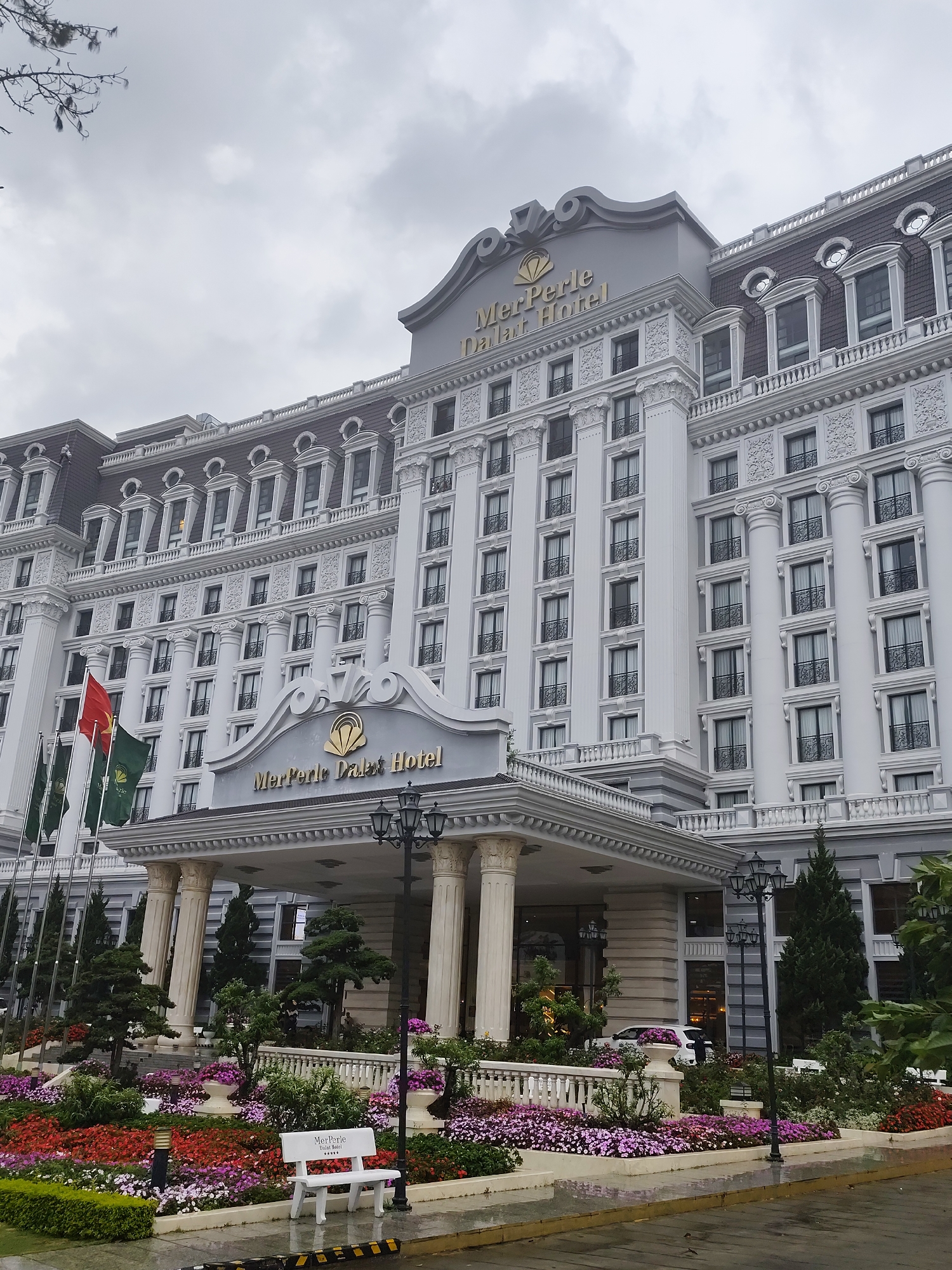 The image shows the front of the MerPerle Dalat Hotel, located in Da Lat, Vietnam. It is a large, multi-story building with a light gray exterior, designed in a neoclassical style. The building has numerous windows, columns, and ornate architectural details. Flags are flying in front of the hotel to the left of the entryway. A garden with colorful flowers and various plants adorns the foreground. A white bench with the hotel's name and star rating is visible among the plants. Street lights are positioned around the entrance. The sky is overcast. A white car is parked under the canopy that is at the entrance. The ground appears wet, likely from rain.