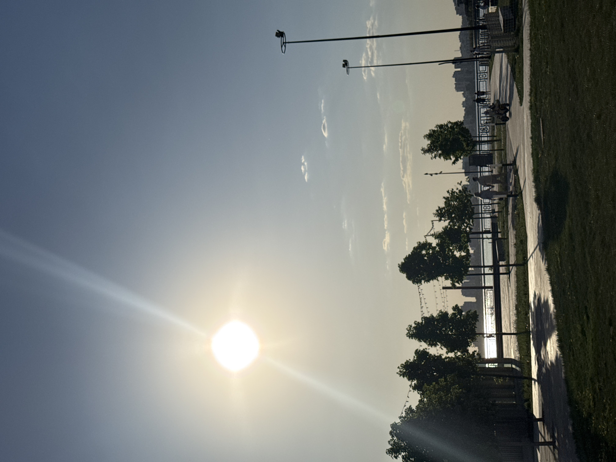 The image depicts a sunny scene along a waterfront promenade in Kazan, Russia. The sun is positioned low in the sky, creating a strong glare. Along the promenade, there are trees, benches, and decorative railings. Several people are visible in the distance, appearing to be walking or riding scooters. Lampposts are spaced along the walkway. In the background, across the water, is the skyline of Kazan. The promenade is bordered by a grassy area.