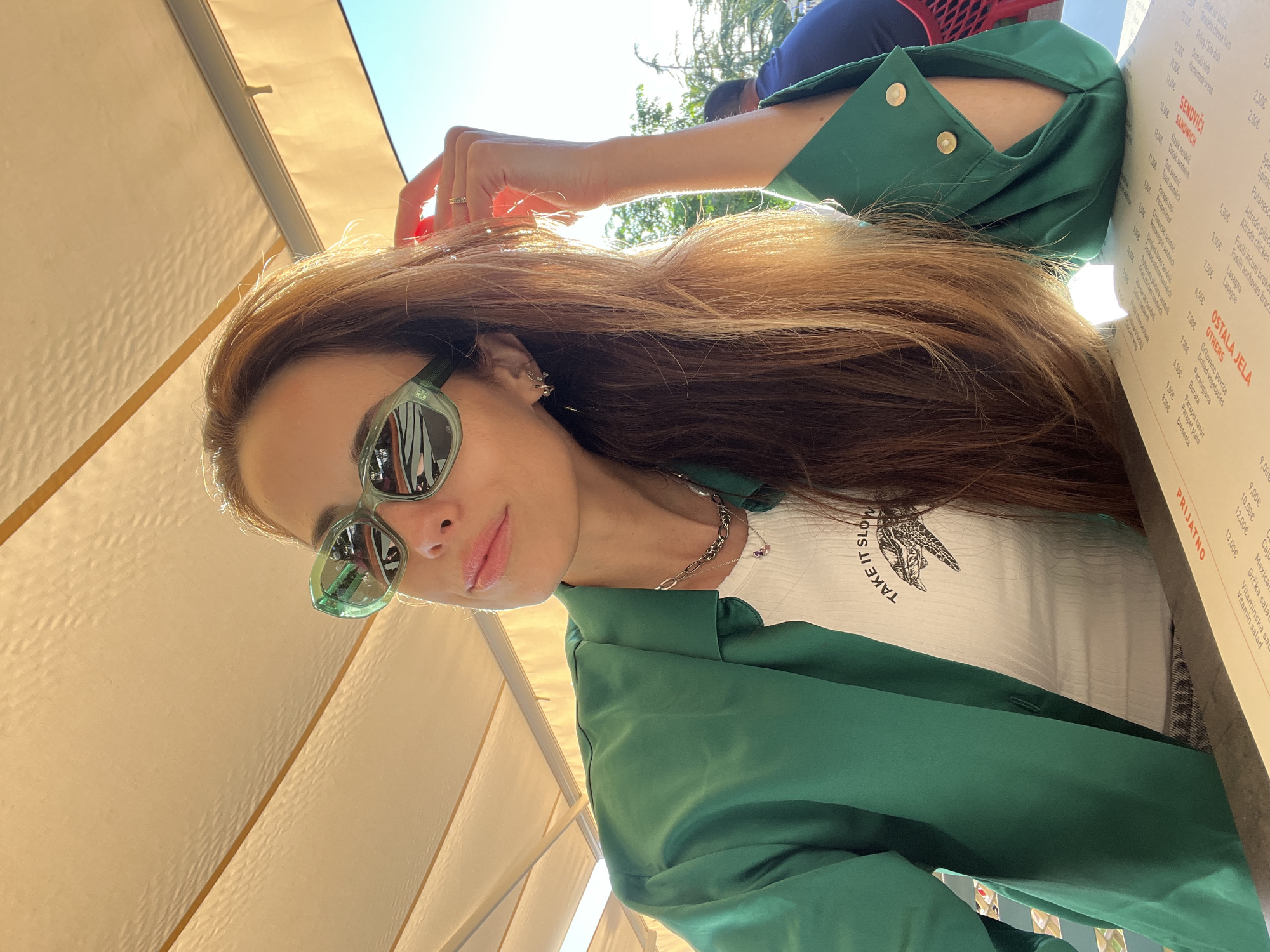 An outdoor, tilted eye-level shot captures a young woman posing at what appears to be a sunny cafe in Bar, Montenegro. She looks directly at the camera with a slight smile, wearing green-framed sunglasses that reflect the surroundings. Her long, light brown hair, illuminated by sunlight, cascades over her shoulders. She is dressed in a vibrant green jacket or blouse, worn open over a white, ribbed t-shirt featuring a graphic animal (possibly a turtle or bird) and the text "TAKE IT SLOW". Delicate earrings, a layered necklace with a chain and small purple stones, and a ring on her right hand are visible accessories.
The setting suggests an outdoor dining area, possibly under a light-colored awning or umbrella, providing shade. Bright sunlight streams from above and behind, creating highlights on her hair and a warm glow on the scene. A person in a blue top is partially visible in the blurred background. Next to the woman, a menu from the establishment is prominent, listing various food items like "SENDVIČ" (SANDWICH), "Grilovano povrće" (Grilled vegetables), "Parmigiana", "Lasagna", and different salads, with prices in Euros. The word "PRIJATNO" (Enjoy) is also seen at the top of the menu. The overall atmosphere is relaxed and casual, characteristic of a daytime meal or break.