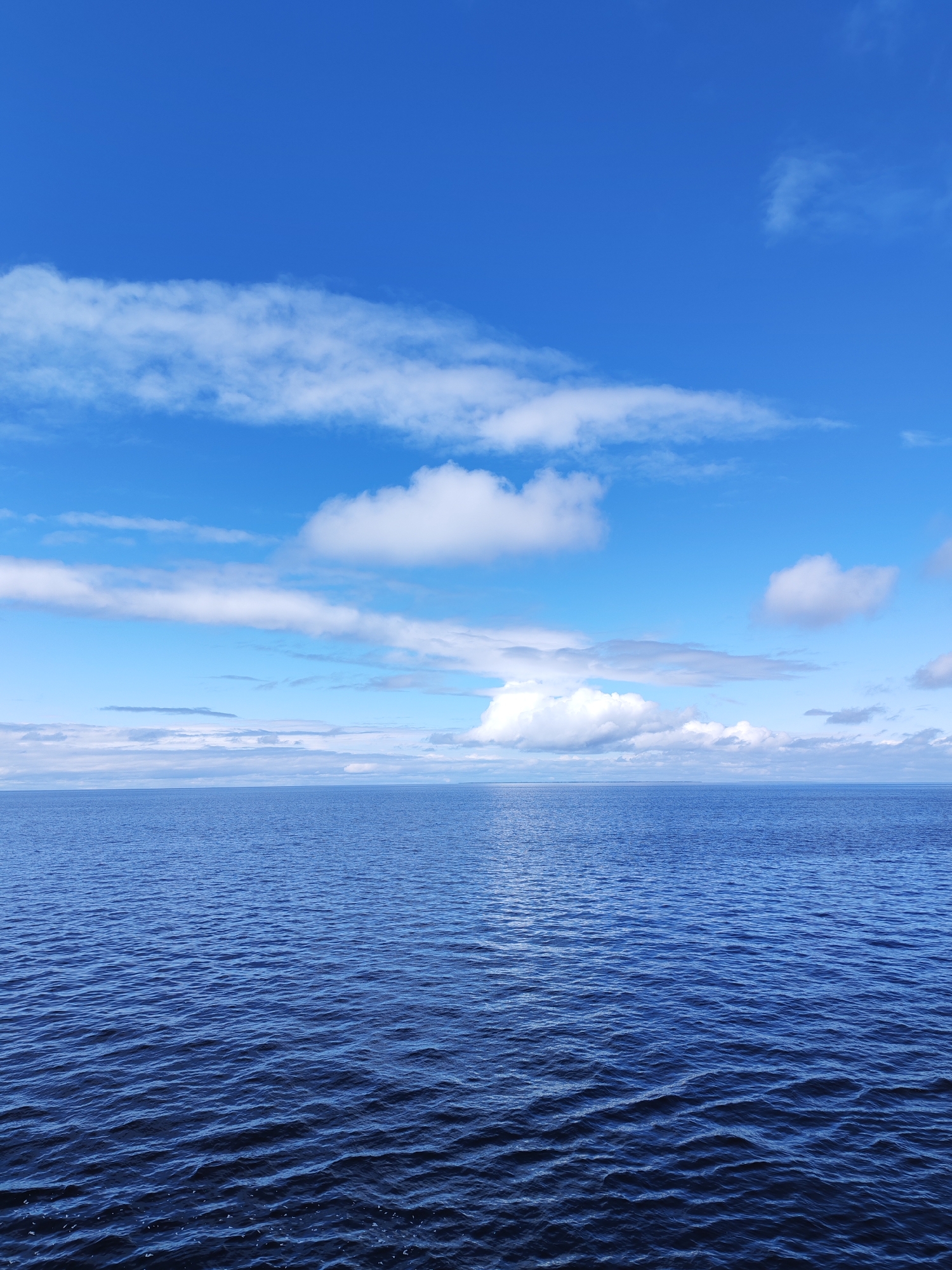 The image depicts a body of water under a bright blue sky with scattered white clouds. The water is a deep blue color with visible ripples and wave patterns. The horizon line is visible in the distance, with a strip of land or possibly more clouds on the horizon. No people or distinct objects are present. The location is unknown, but presumed to be in Unknown, Russia.