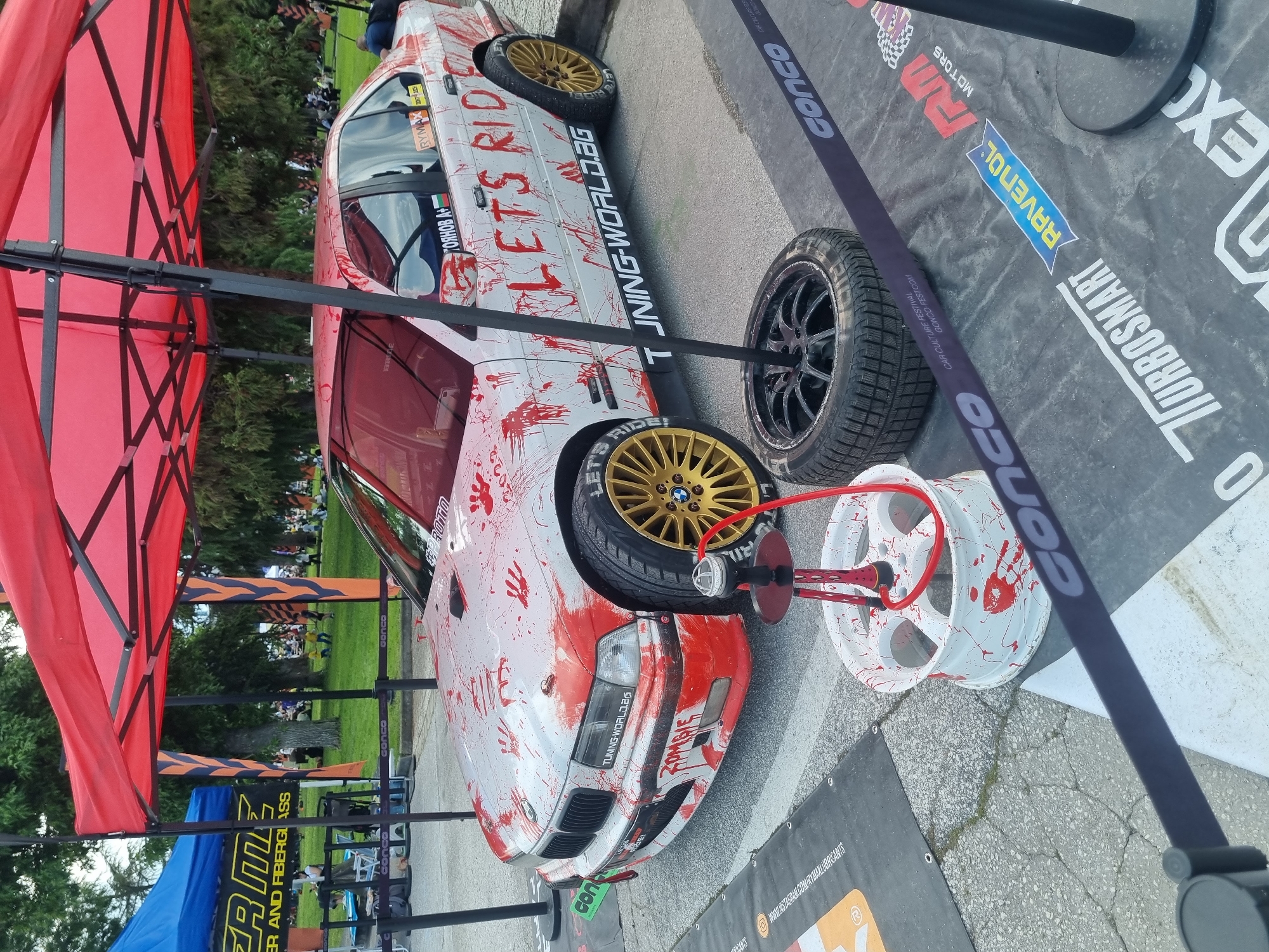 The image features a customized car displayed at a car culture festival, likely in Plovdiv, Bulgaria. The car is white and decorated with what appears to be red paint simulating blood splatters and handprints, giving it a zombie-like theme, including the words "ZOMBIE" on the front bumper. It also has the words "LET'S RIDE" and "TUNING-WORLD.BG" painted on the side, along with the name "TOЯНОВ А." (Toyanov A) and the Bulgarian flag. The car has gold-colored wheels, and additional wheels, one white with red splatters and another black, are placed beside it. A hookah pipe sits next to the white wheel. The car is parked on an asphalt surface covered with marketing banners featuring logos and brand names such as "TURBOSMART," "RAVENOL," and "CONCO," which are likely sponsors of the event. A red canopy is partially visible in the foreground. The event seems to be held on a grassy area, with people visible in the background. Overall, the scene conveys a customized car display at a car culture festival with a distinctive zombie theme.