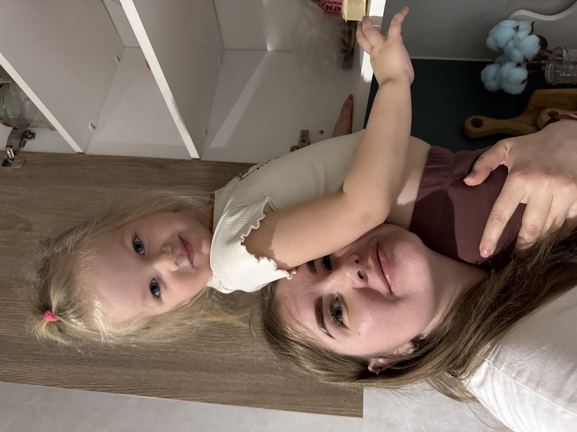 A woman and a child are lying down in a room in Gornyi (Dvurechenskoe S/P), Russia. The child, a young girl with blonde hair tied back with a pink hair tie, is lying with her head on a wooden surface. The woman, who has blonde and brown hair, is lying next to her on a white pillow. The girl is wearing a white shirt, and the woman is wearing a white shirt and a brown top. They are positioned next to a white kitchen cabinet with items inside.
