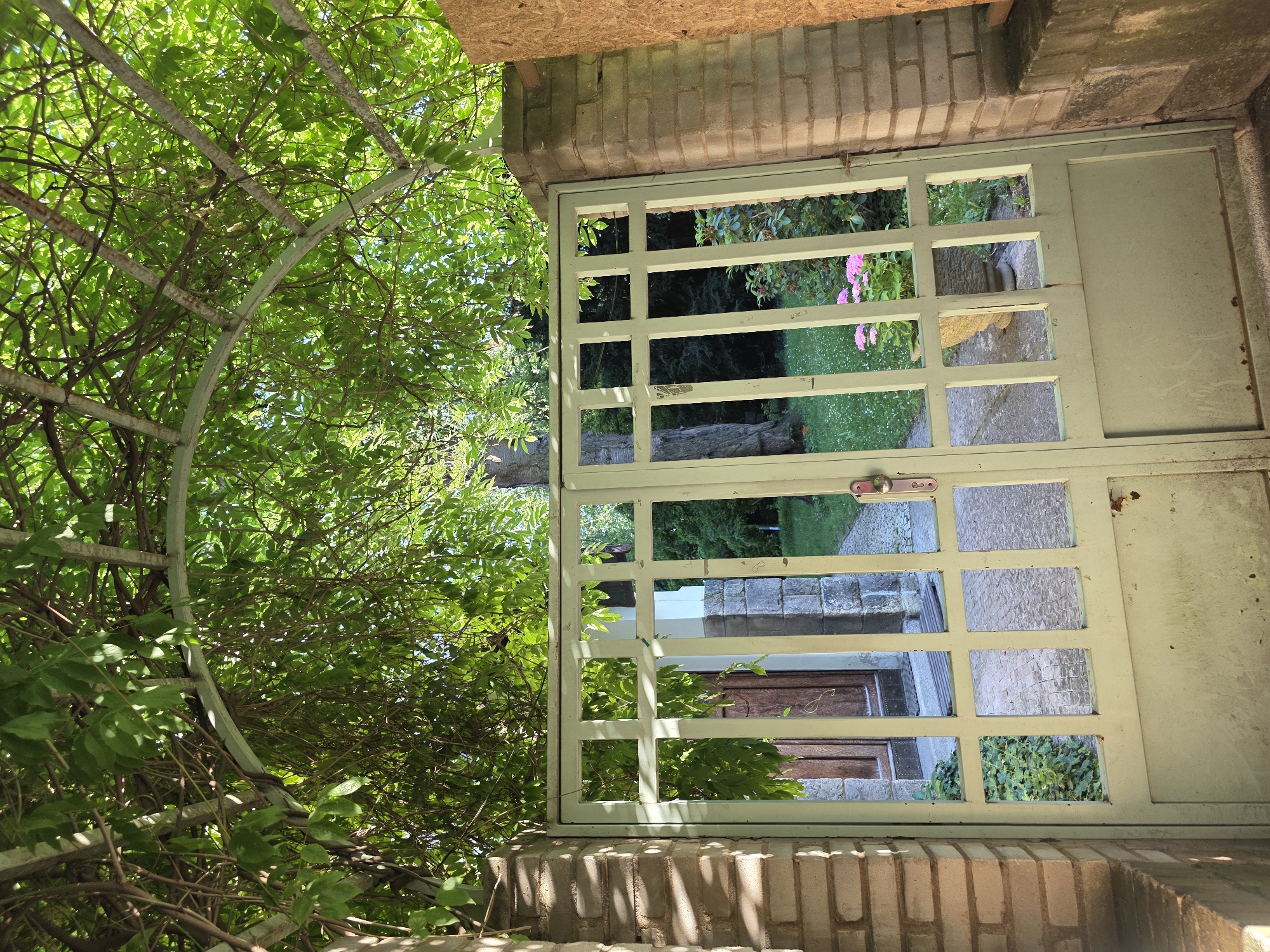This image captures a view through a light green metal gate with a grid pattern. The gate is set into a brick wall, which is topped with a wooden beam and a stone facade. Lush green foliage, likely from a vine or tree, drapes over a curved metal arbor to the left of the gate.
Through the gate's openings, a garden path with stone steps is visible, leading towards a wooden door. In the background, there are more trees and hints of pink flowers. The scene appears to be outdoors during daylight, with sunlight filtering through the leaves and casting shadows on the brickwork. The overall impression is one of an overgrown, serene garden entrance. There is no discernible text in the image.
