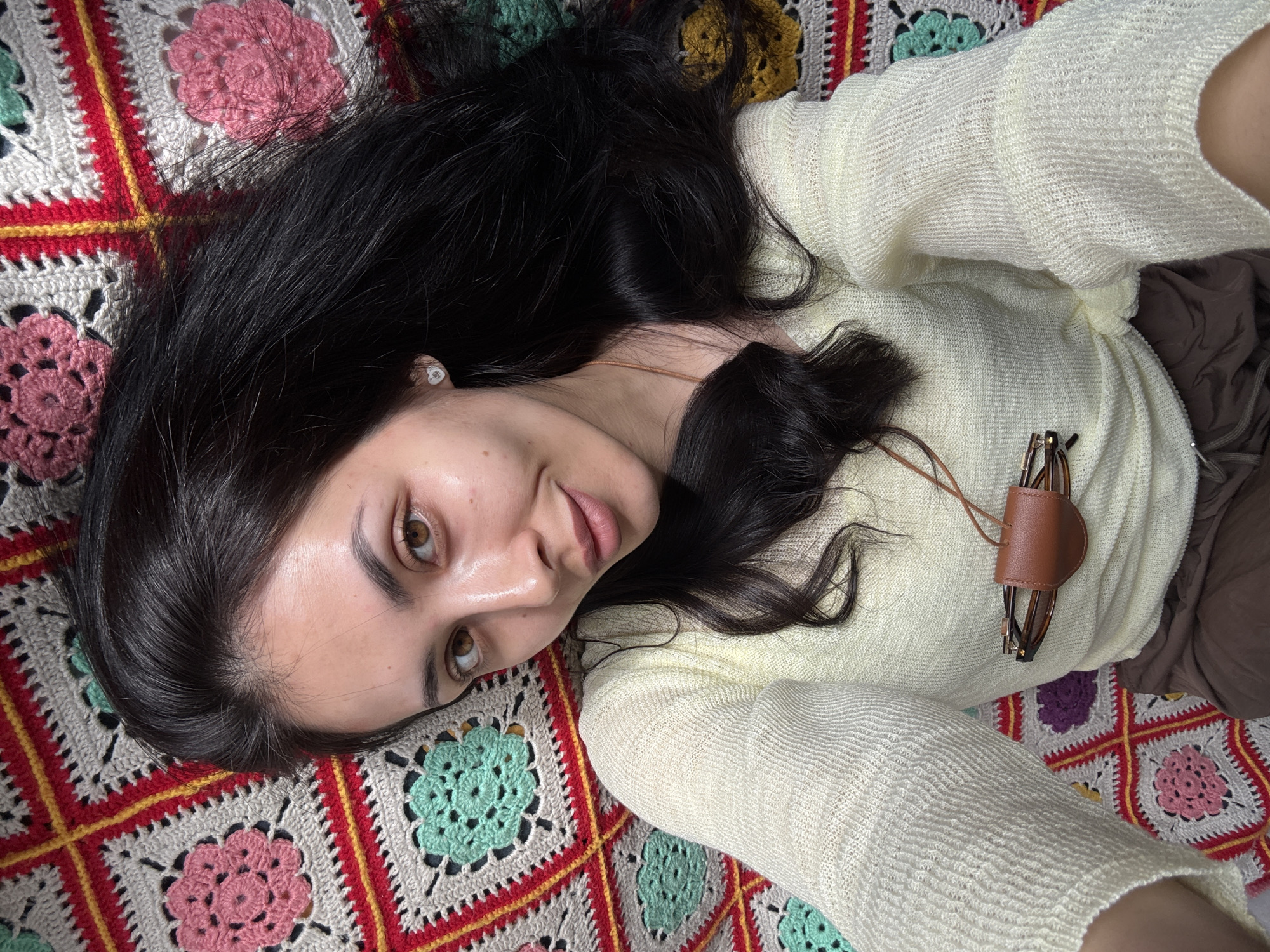 A young woman with long, dark hair and light brown eyes is depicted from an overhead perspective, lying on a vibrant, crocheted blanket. She is looking upwards, directly at the camera, with a calm and gentle expression, featuring a slight smile. Her hair is spread out around her head, and a small, light-colored stud earring is visible in her left ear.
She is dressed in a light yellow or cream-colored, long-sleeved top, made of a textured, possibly knitted or loosely woven, fabric. A brown leather holder with a pair of brown-framed glasses hangs from a cord around her neck. The lower right portion of the image shows a glimpse of brown fabric, likely part of her attire.
The background is a colorful crocheted blanket composed of granny squares. These squares feature central floral motifs in shades of pink, mint green, and yellow-gold, enclosed by borders of white, red, and yellow yarn, creating a rich and intricate pattern. The overall setting suggests a comfortable, indoor environment. The image primarily focuses on the woman's face and upper body against the textured backdrop. No specific time of day or weather can be determined from the visual information. There is no visible text in the image. The external context indicates the location is Votkinsk, Russia.