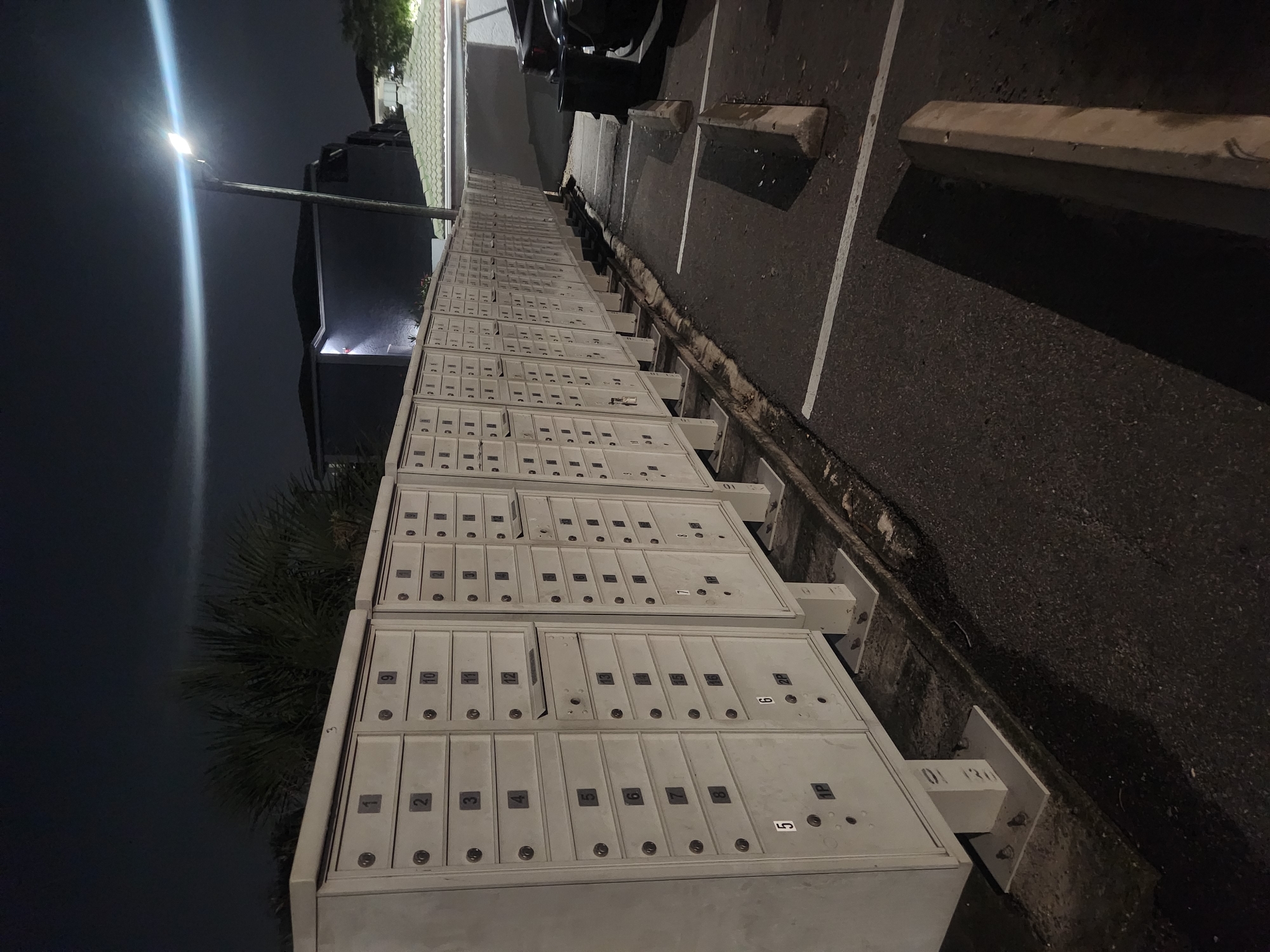 A long row of outdoor mailboxes stands in a dimly lit parking lot at night. The mailboxes are arranged in columns and rows, with each individual mailbox clearly numbered. Some mailboxes have visible numbers like 1, 2, 3, 4, 5, 6, 7, 8, 9, 10, 11, 12, 13, 15, and 16. A street light illuminates the scene, casting long shadows and creating a bright glare. In the background, there are apartment buildings and trees, including palm trees. The parking lot has painted white lines and concrete wheel stops. A black trash can and parts of vehicles are also visible. The ground is asphalt. The image appears to be taken in Altamonte Springs, Florida, USA.