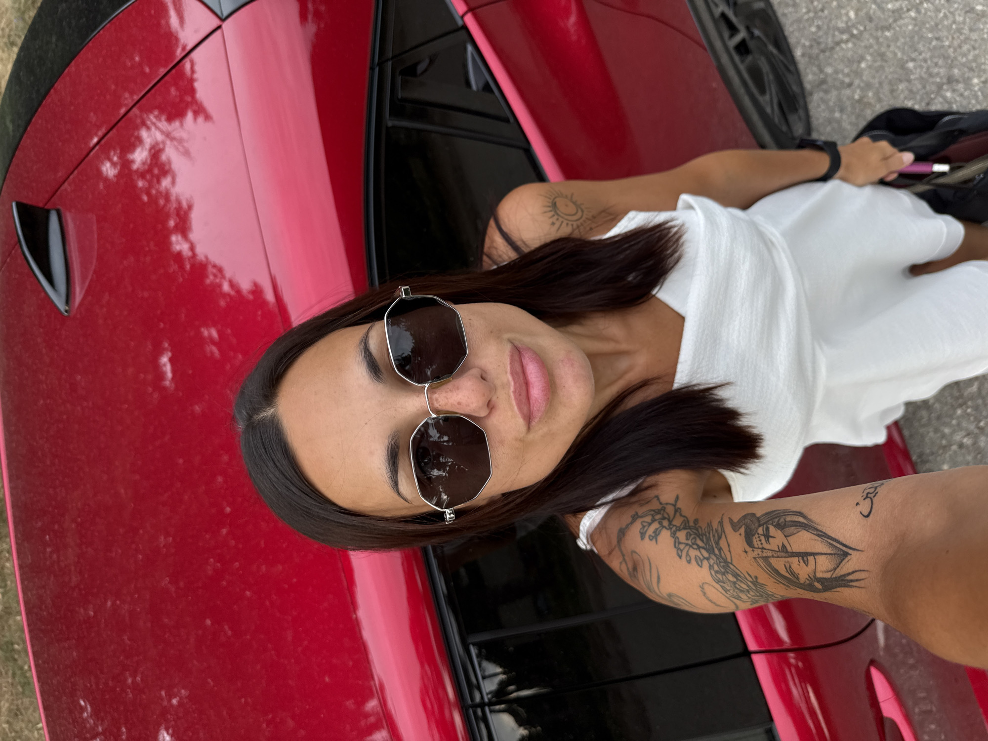 A young adult woman stands next to a red car. She is wearing a white dress and sunglasses. Her left arm is visible, adorned with tattoos. The woman is holding a black bag in her right hand. The scene appears to be outdoors, with asphalt visible on the ground. There is no specific indication of the location being in the city of Pleven, Bulgaria.