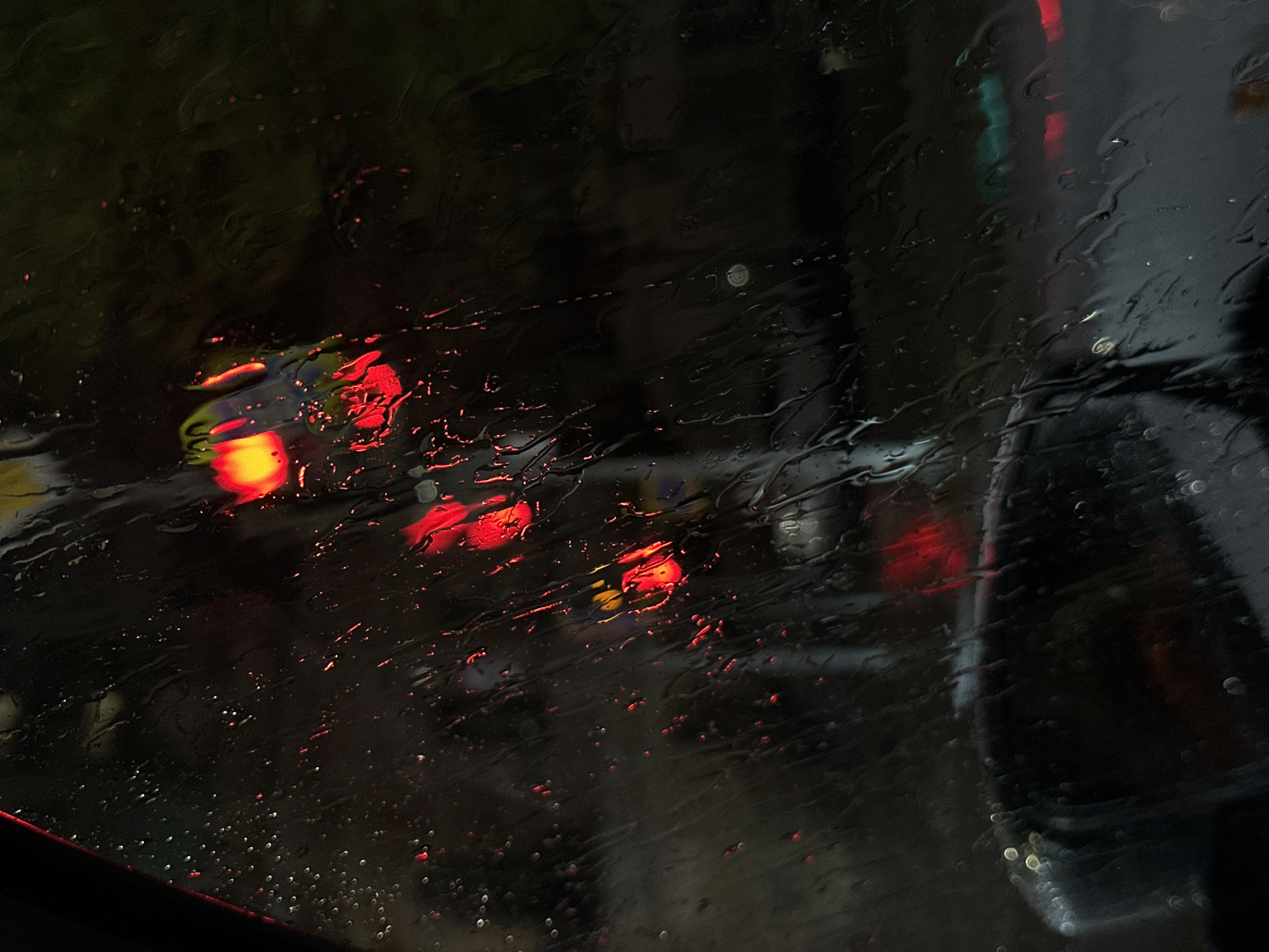 The image appears to be taken from inside a vehicle on a rainy night, focusing on the rain-streaked windshield. The dominant colors are dark shades with blurred and distorted reflections of traffic lights (primarily red and yellow) and other vehicles. The exterior is indistinct. There is no information in the image to determine a specific location in the city of Ovoshchnoy, Russia or to identify people, subjects, or actions within the scene.