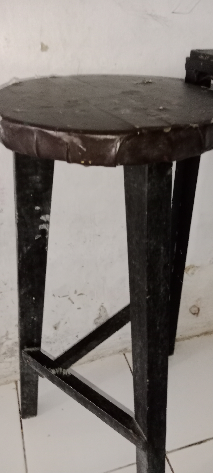 The image shows a dark, circular stool with a dark wooden top and dark metal legs. The stool has a footrest connecting two of the legs. The background appears to be a white wall with some scuff marks and a tiled floor. The image is a close-up shot, focusing on the stool. It is not possible to determine the specific location within Subang, Indonesia, or any actions or interactions taking place, as the image only depicts a static object in an indoor setting.