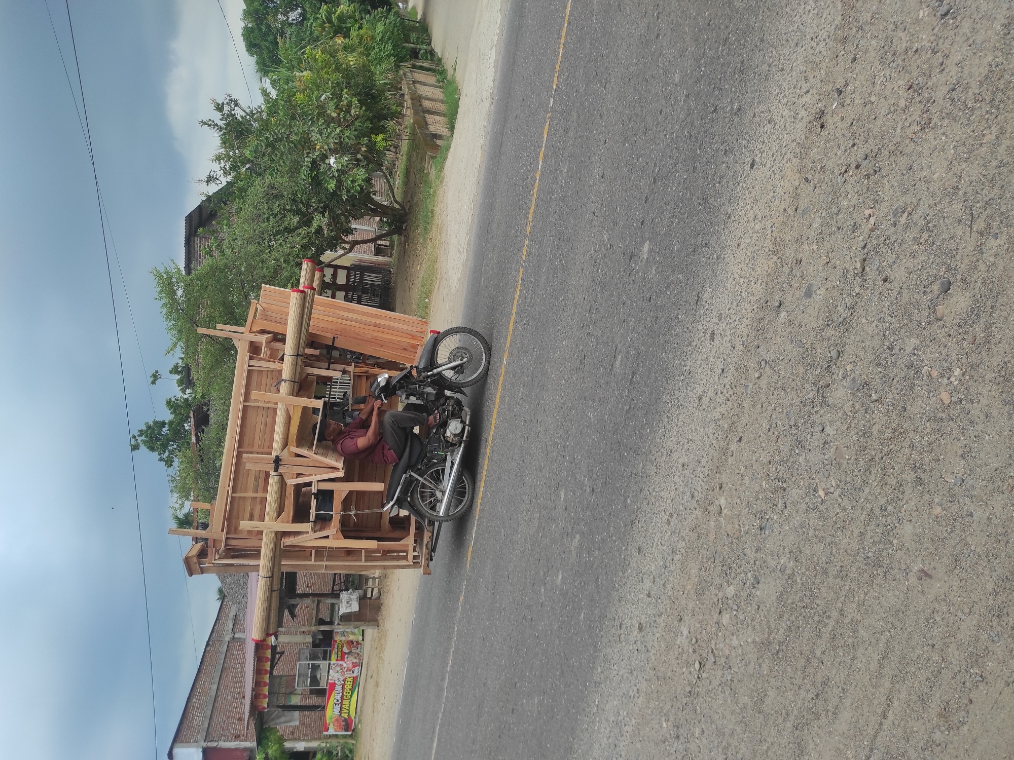 This image, taken in Lancang Barat, Indonesia, captures a man on a motorcycle transporting a large wooden structure along a paved road. The photograph is oriented vertically, with the road running horizontally across the right side of the frame and the sky dominating the left.
The central subject is a man, dressed in a red-brown shirt and dark trousers, seated on a dark-colored motorcycle. Attached to the motorcycle is an elaborate, light-toned wooden framework, extending significantly around the vehicle, suggesting it's designed to carry a substantial load or serve as a mobile stall. Bundles of rolled materials, possibly mats or thin wooden poles, are secured to the top of this wooden structure with red and white ties.
In the background, on the side of the road, are several low-rise buildings, some constructed from red brick, partially visible among abundant green trees and foliage. Overhead, thin power lines stretch across the clear sky, which features scattered white clouds. The asphalt road has a faded yellow dividing line, and the unpaved shoulder consists of dirt and gravel.
A visible banner or sign attached to one of the background buildings displays text advertising food: "MIE CELOR & AYAM GEPREK," indicating the presence of local food vendors. The scene depicts a common form of resourceful transport and commerce in a rural or semi-urban Indonesian setting, occurring during bright daytime hours.